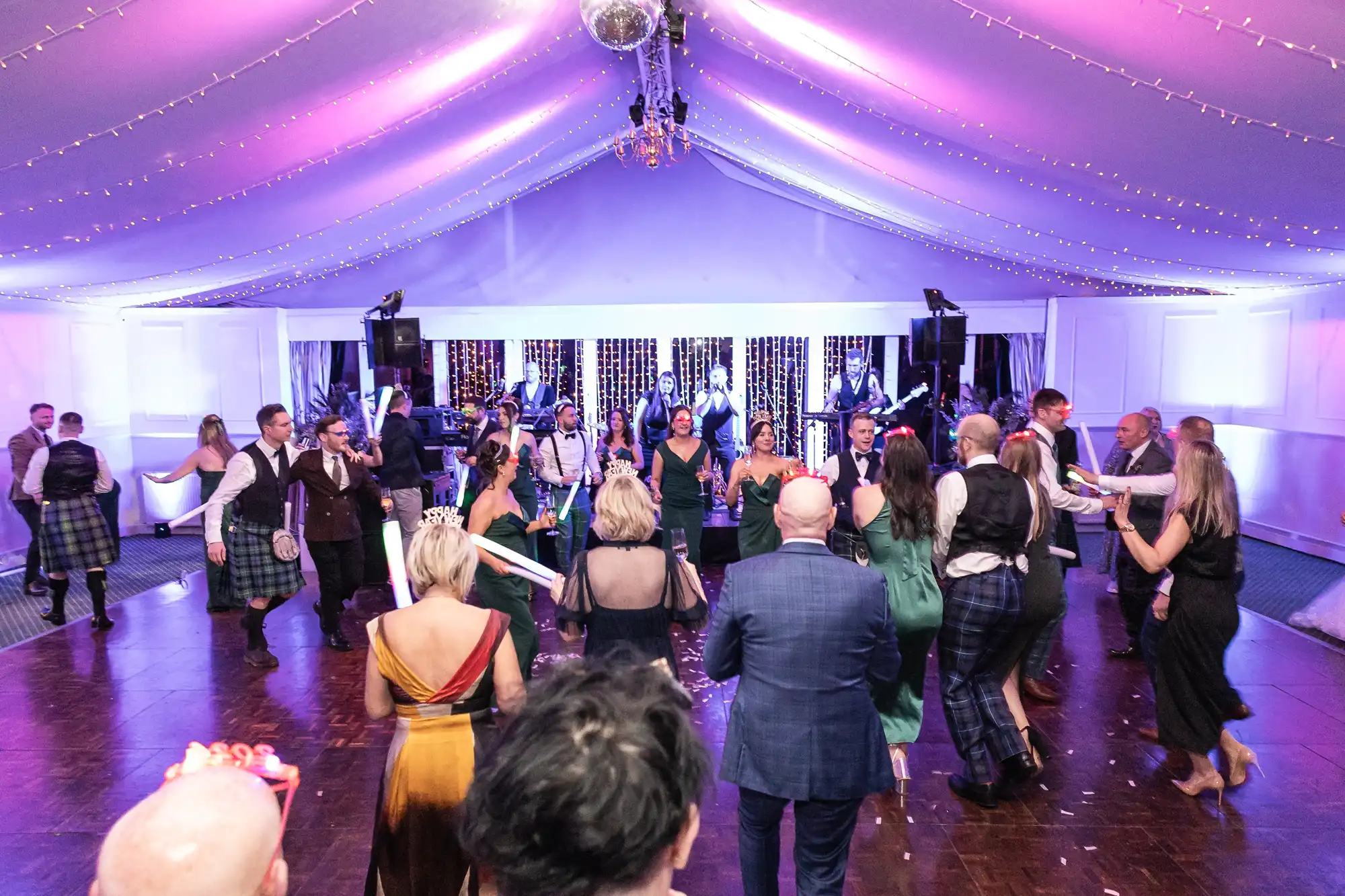 A group of people dance under a tent with purple lighting and a disco ball, while a live band performs on stage in the background.