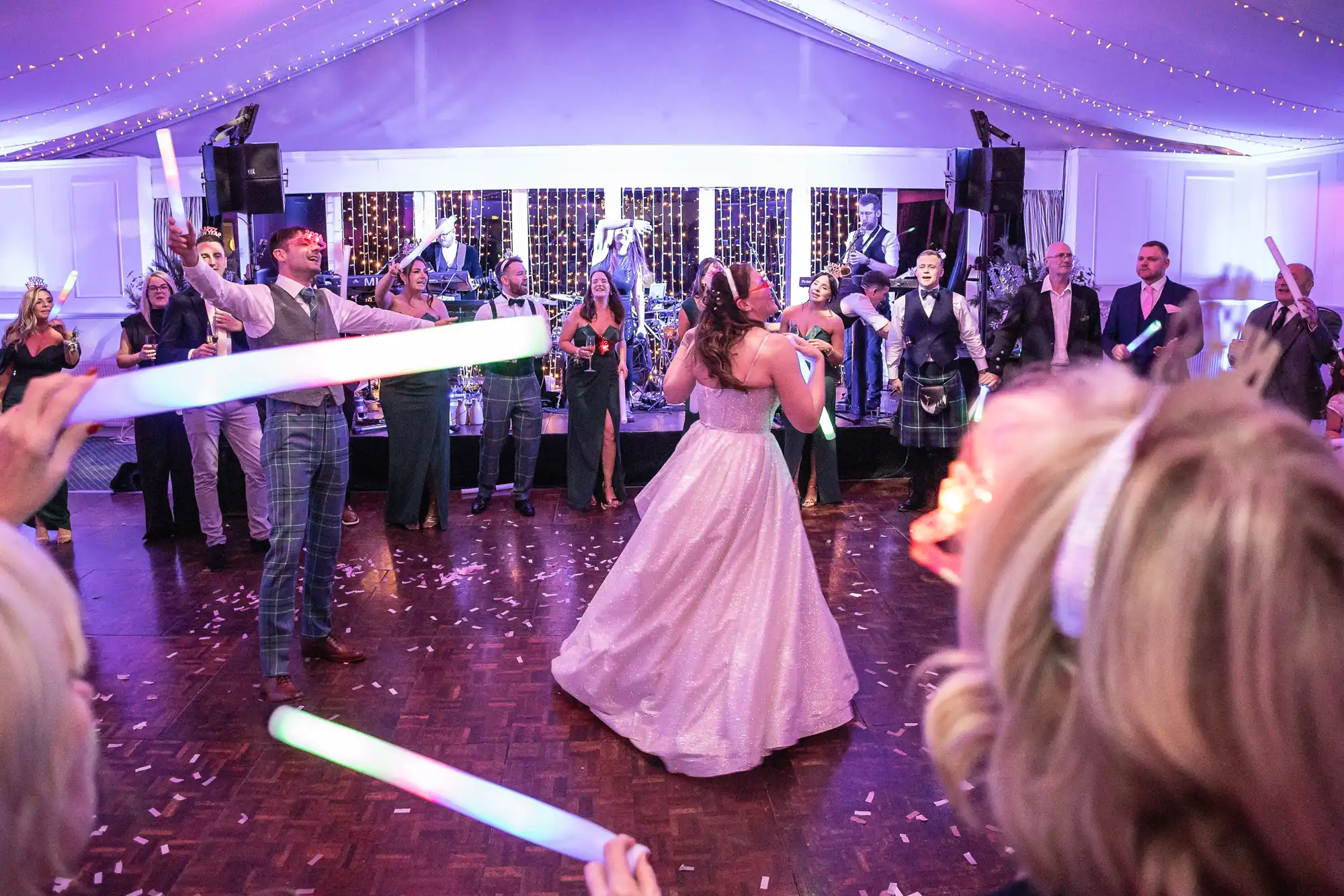 A bride and groom, flanked by their wedding party, dance energetically with light sticks on a dance floor under ceiling drapes illuminated by colorful lights.