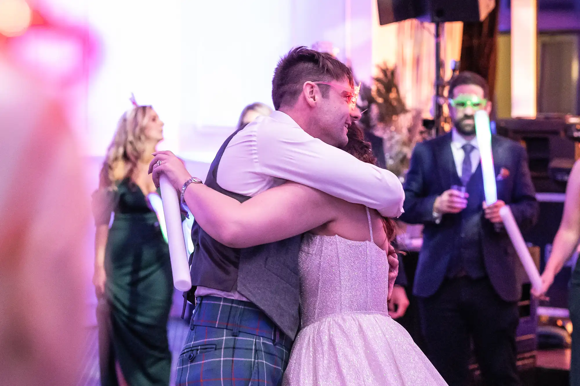 A man and woman embrace at a lively indoor gathering with colorful lighting and people holding glow sticks.