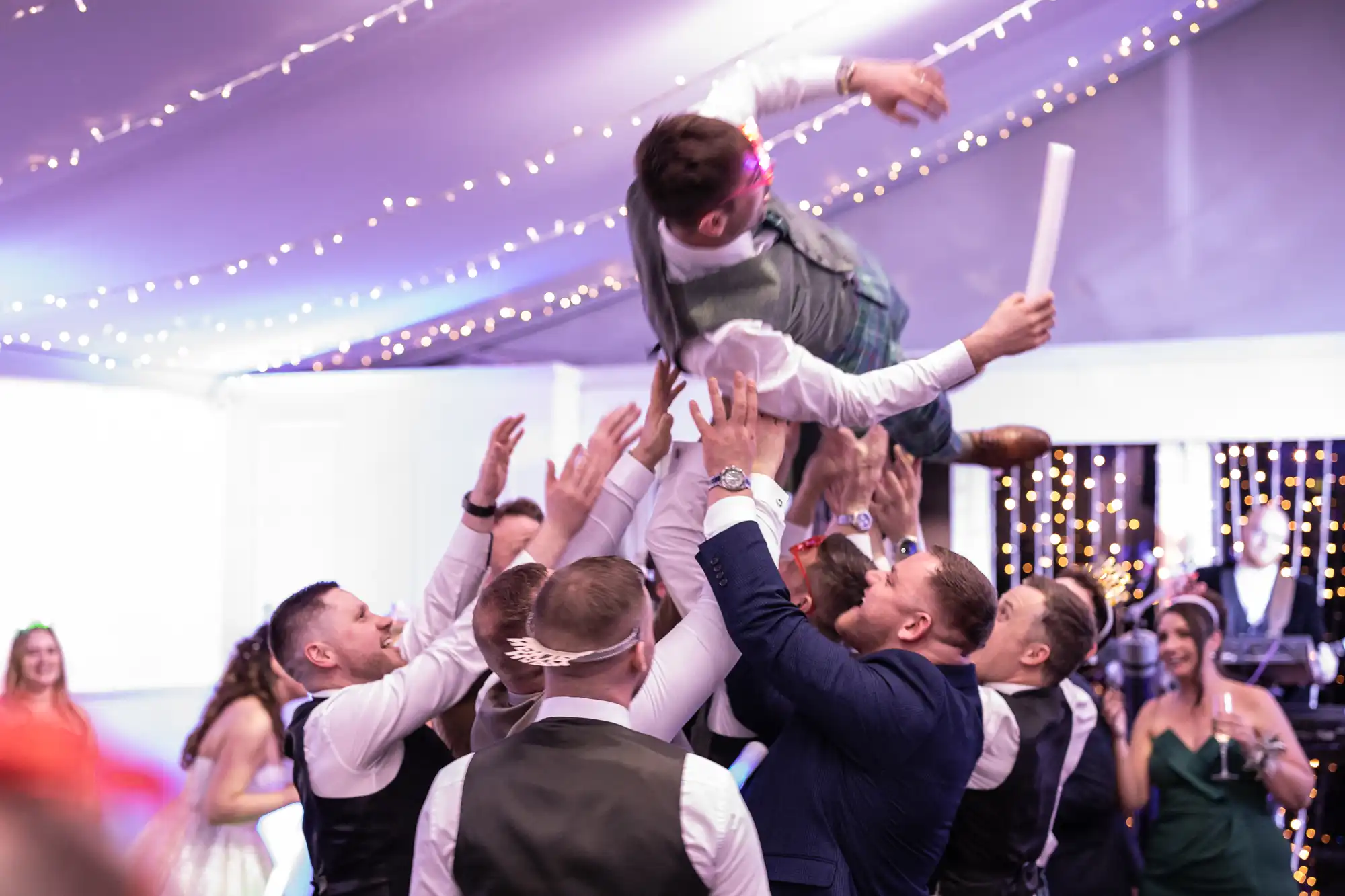 A group of people in formal attire lift a man in the air during a celebratory event with string lights in the background.