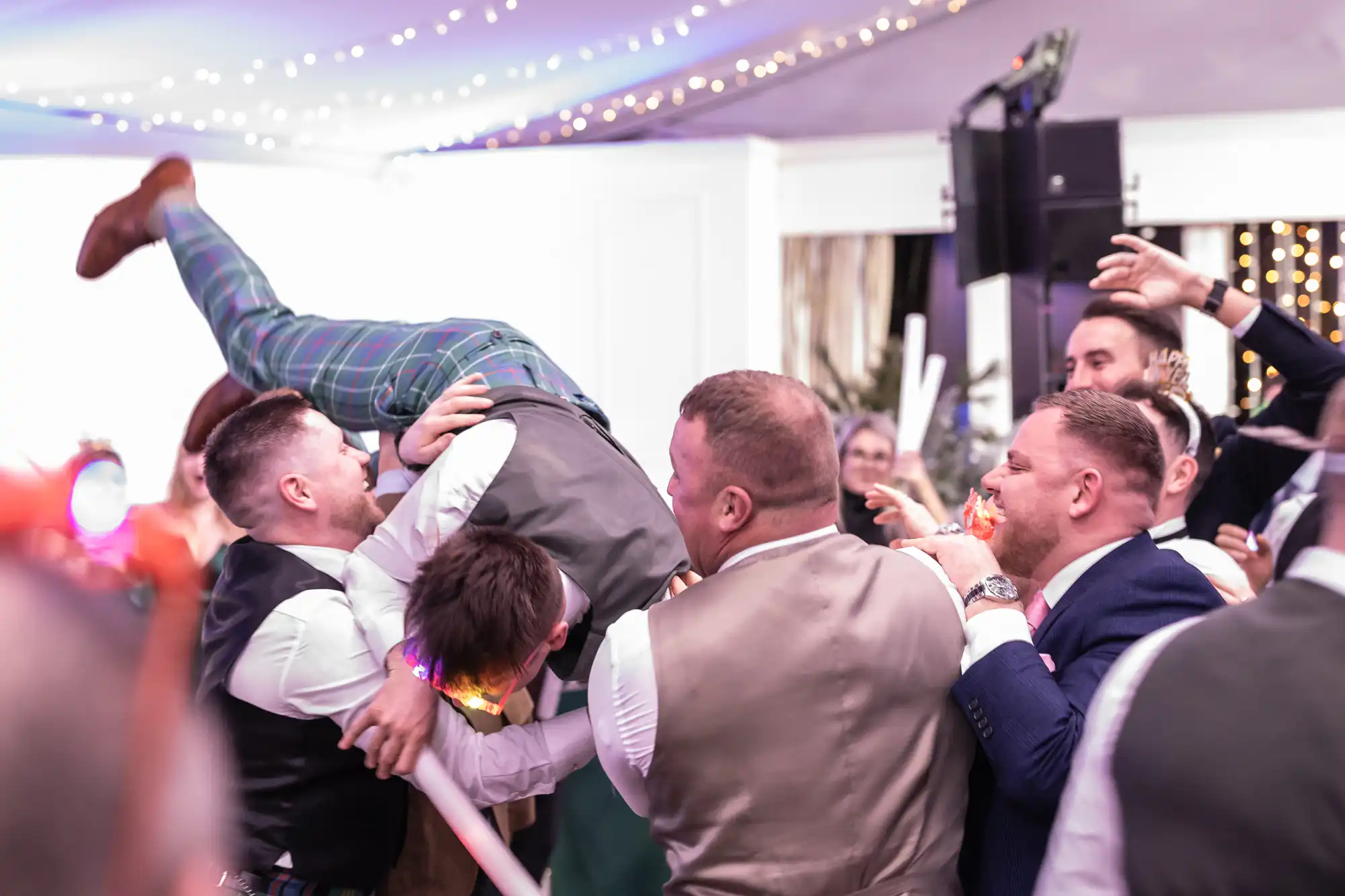 Several men, dressed in vests and suits, are lifting another man upside down during what appears to be a lively celebration or party, with string lights hanging overhead.