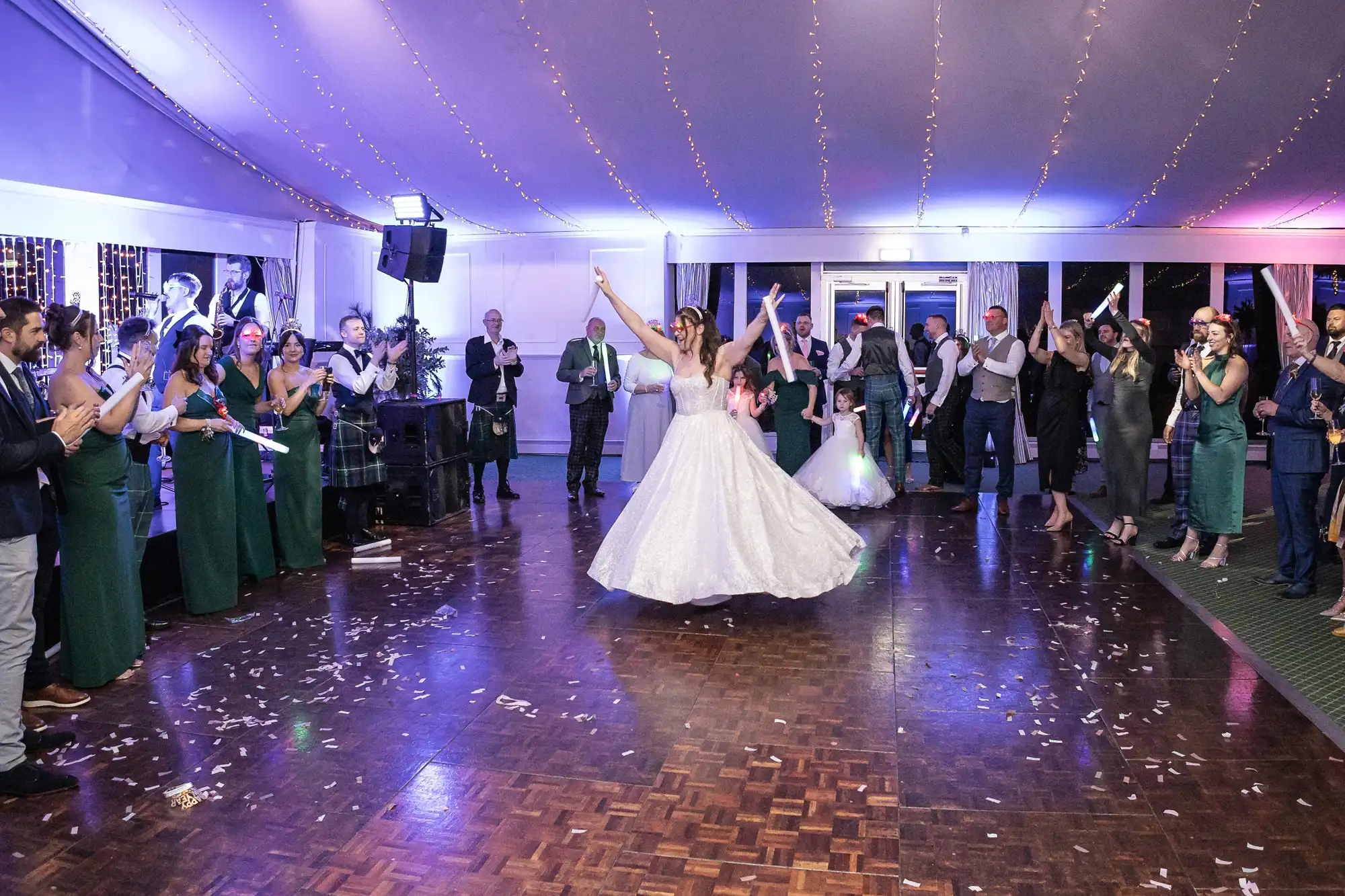 A bride in a white gown dances energetically at her wedding while guests in formal attire watch and cheer inside a decorated event venue.