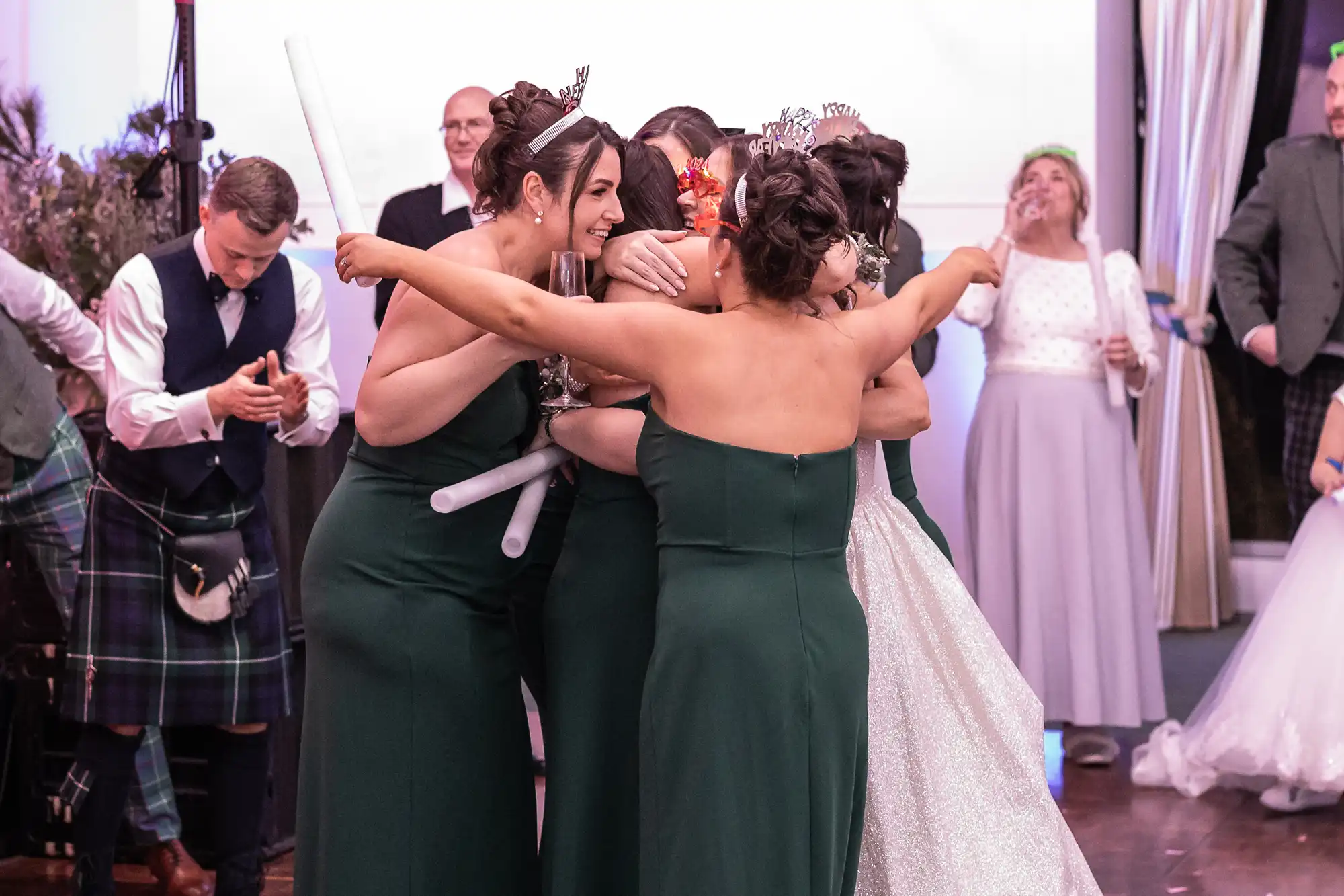 A group of women, dressed in formal attire, share a hug in the center of a room. Other people in similar attire stand nearby, some observing and others engaged in different activities.