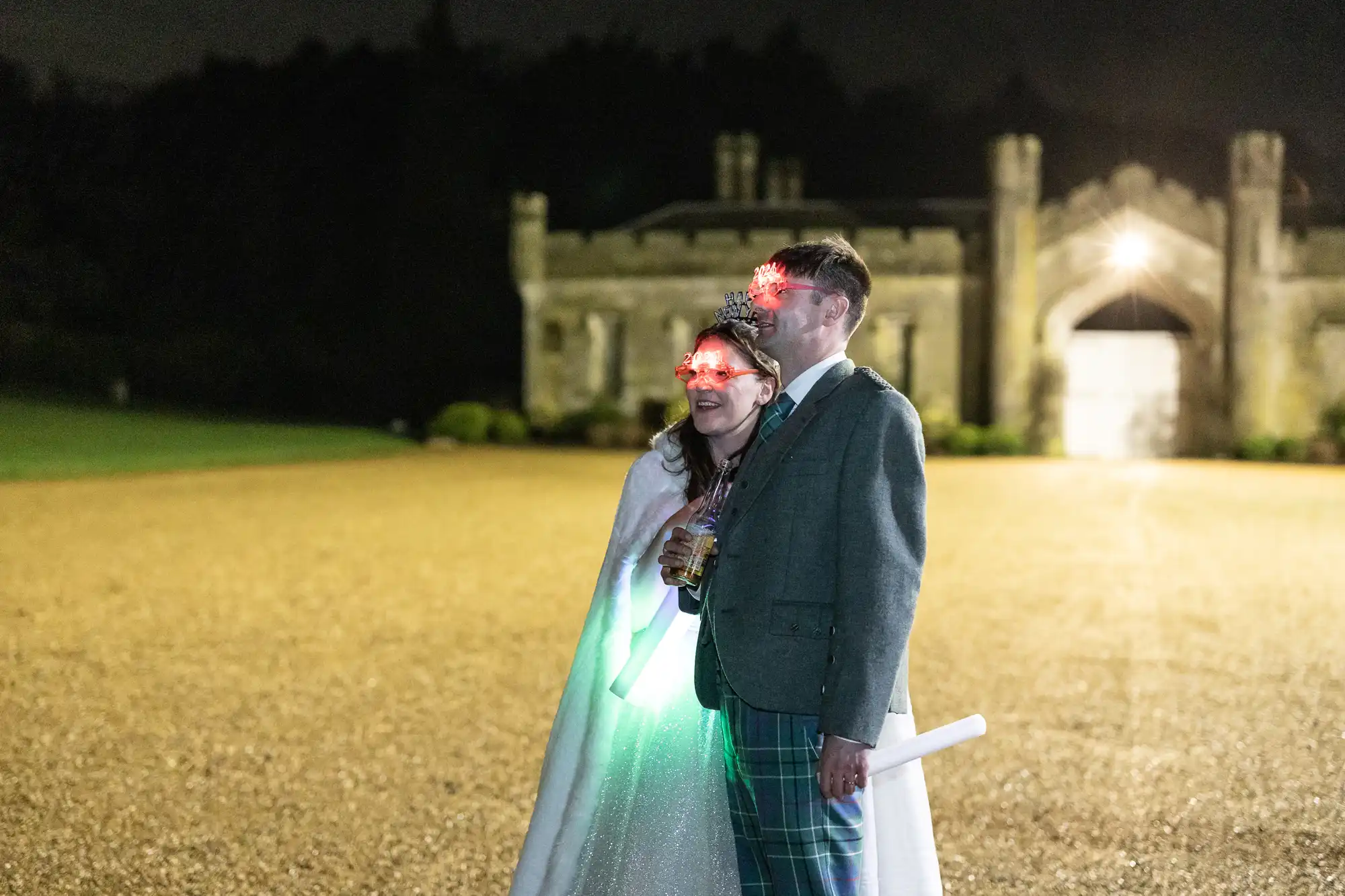 A couple stands outside at night with light-up glasses, dressed in festive attire, including a crown and cape. They hold glowing objects against a backdrop of an illuminated stone building.