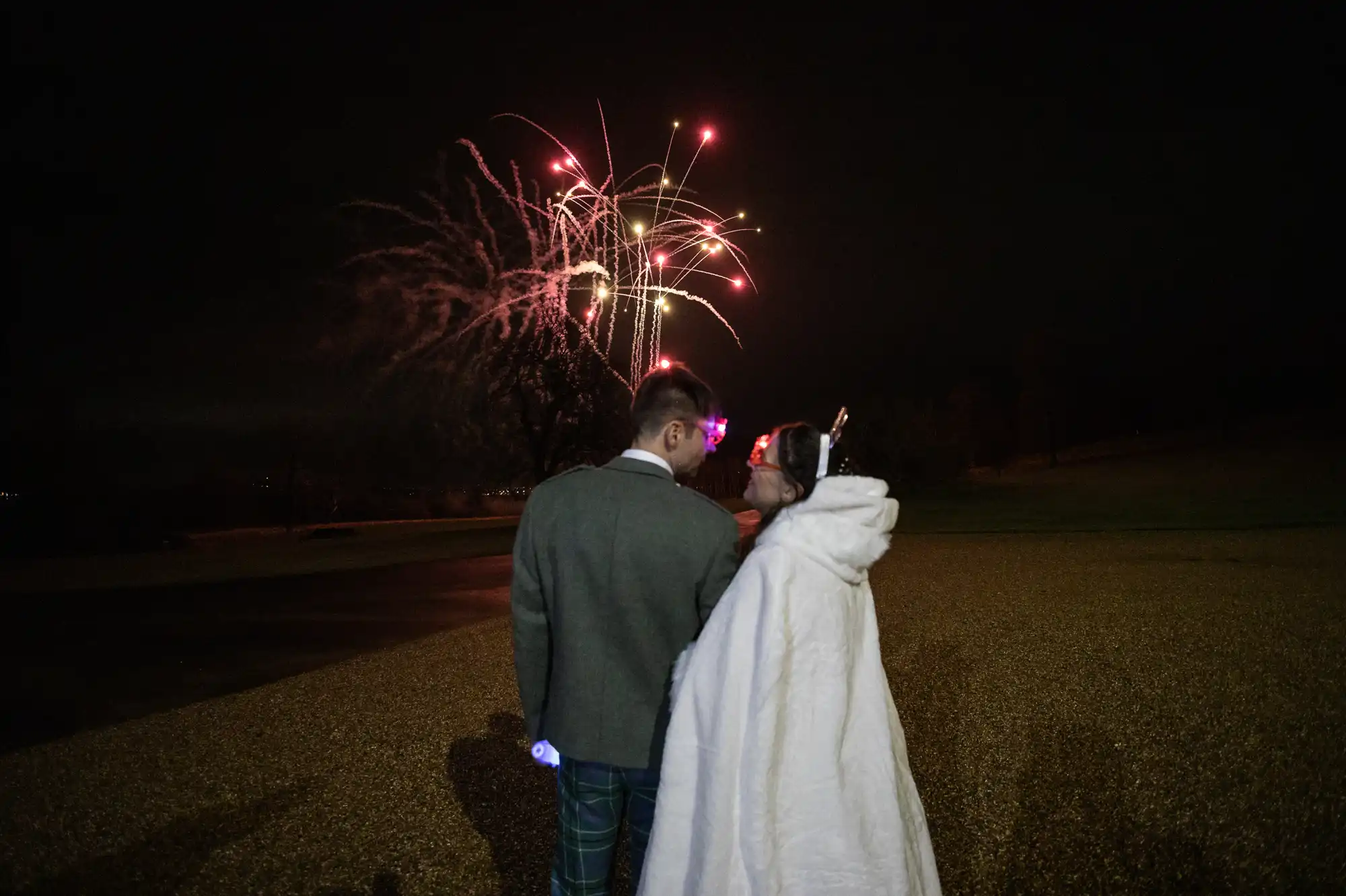 A couple stands in the dark, watching a vibrant firework display in the night sky. The person on the right is draped in a blanket.