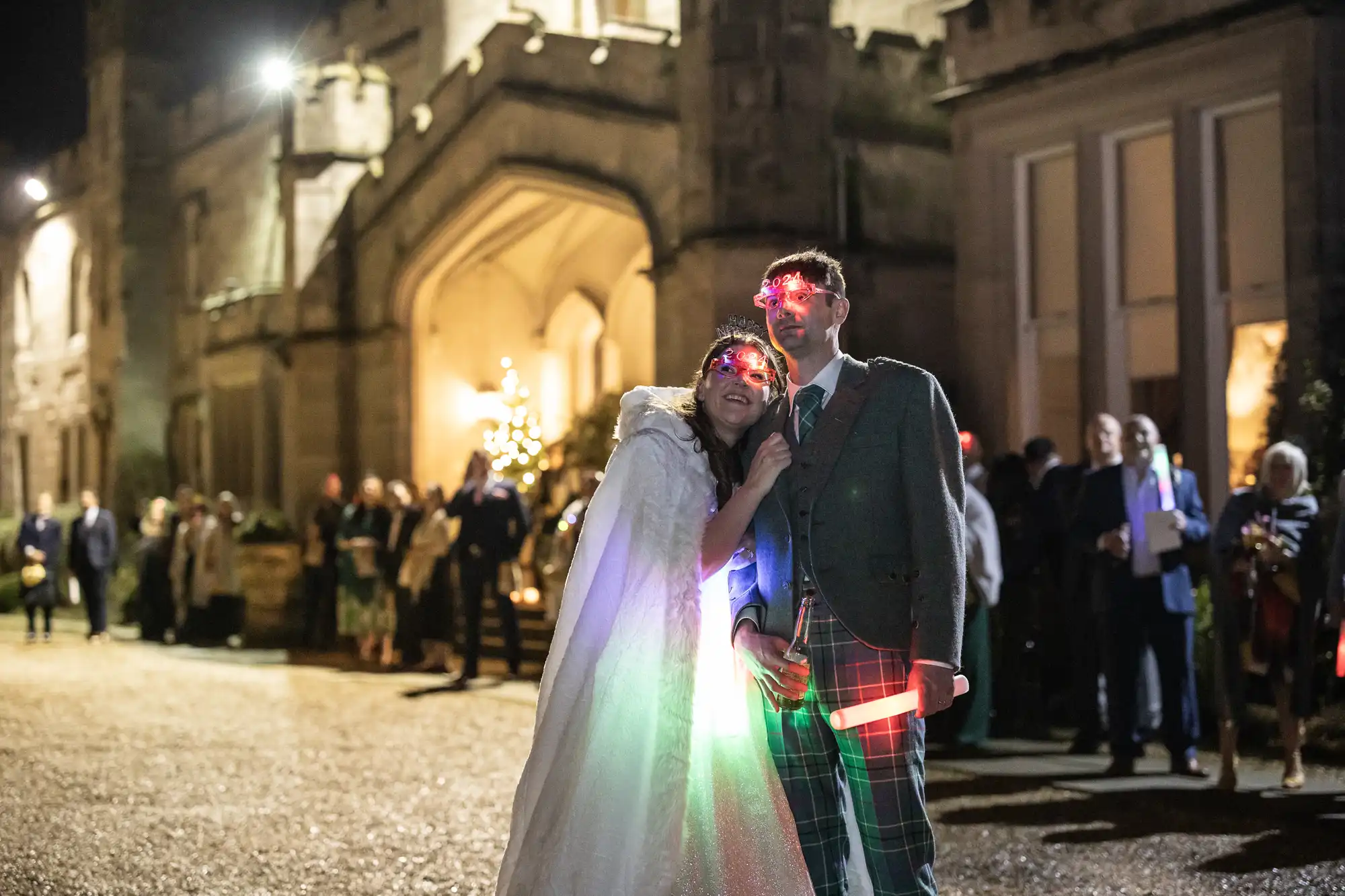 A couple dressed in wedding attire stands outside a lit-up stone building at night, with the bride leaning on the groom. Both hold glowing sticks, and people are gathered in the background.