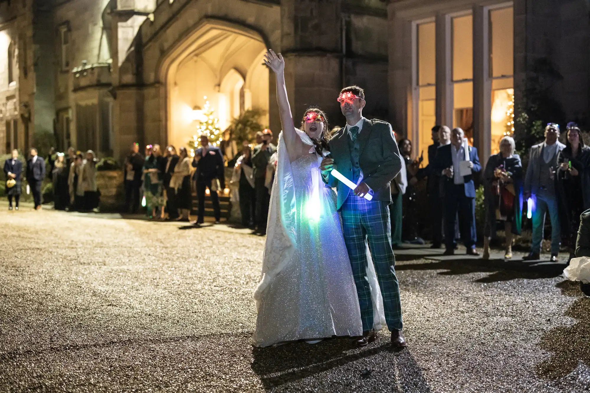 A newlywed couple, illuminated by colorful lights, celebrates outside a large building as a crowd watches in the background. The bride raises her hand joyfully, and both wear novelty glasses.