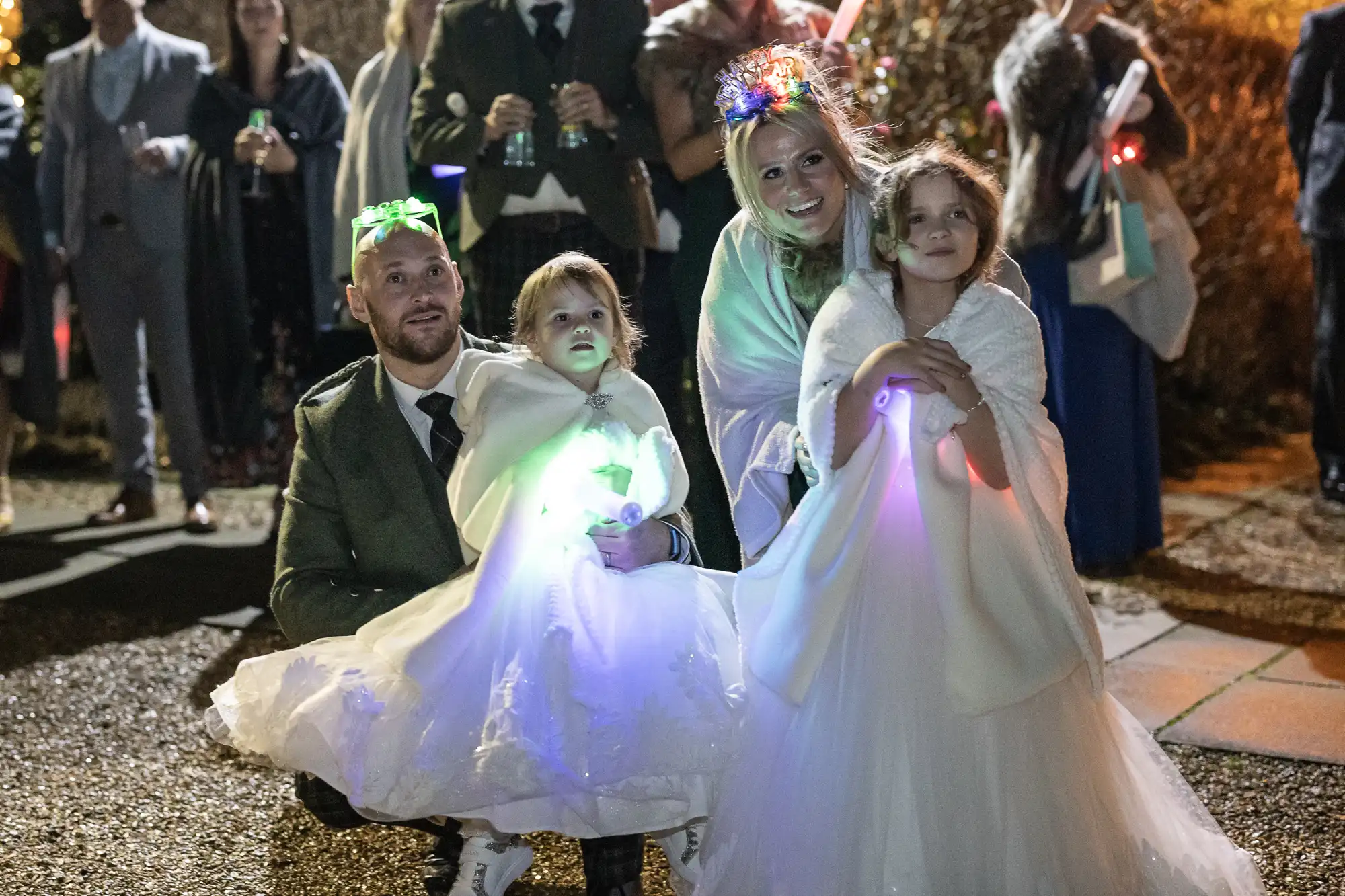 A family dressed in formal attire, with the father and three children wearing white dresses, pose for a photo at an outdoor event. The children hold glowing light-up toys.