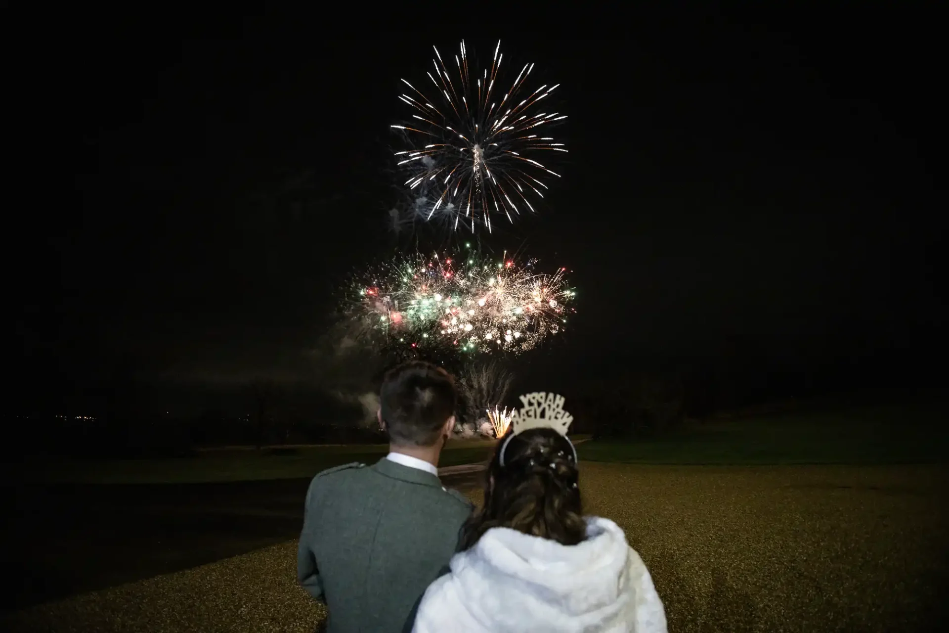 A couple stands facing a nighttime sky filled with fireworks. The woman is wearing a tiara.