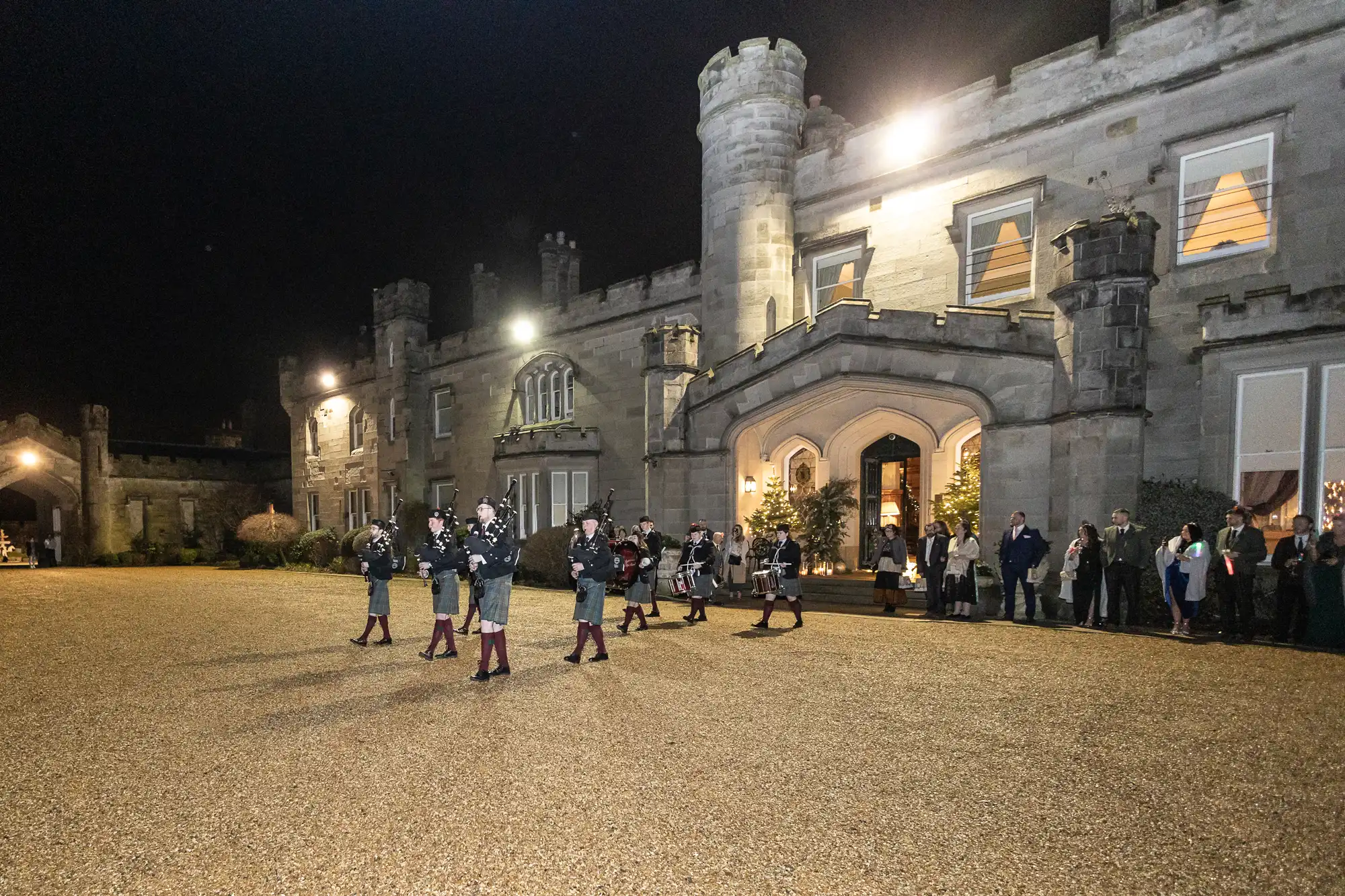 A nighttime scene shows a group of people standing outside an illuminated stone castle with bagpipers performing in front.