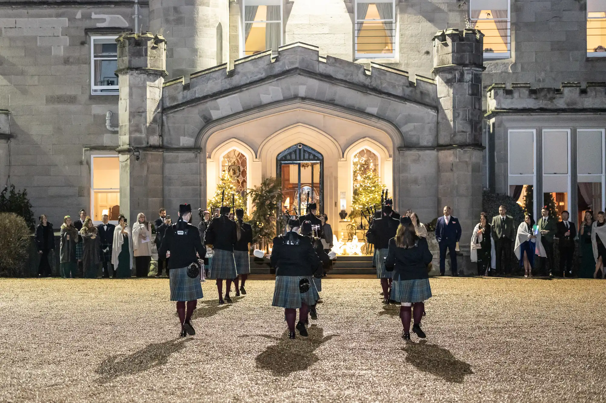 A group of bagpipers in traditional Scottish attire marches toward the entrance of a castle-like building while onlookers stand nearby.