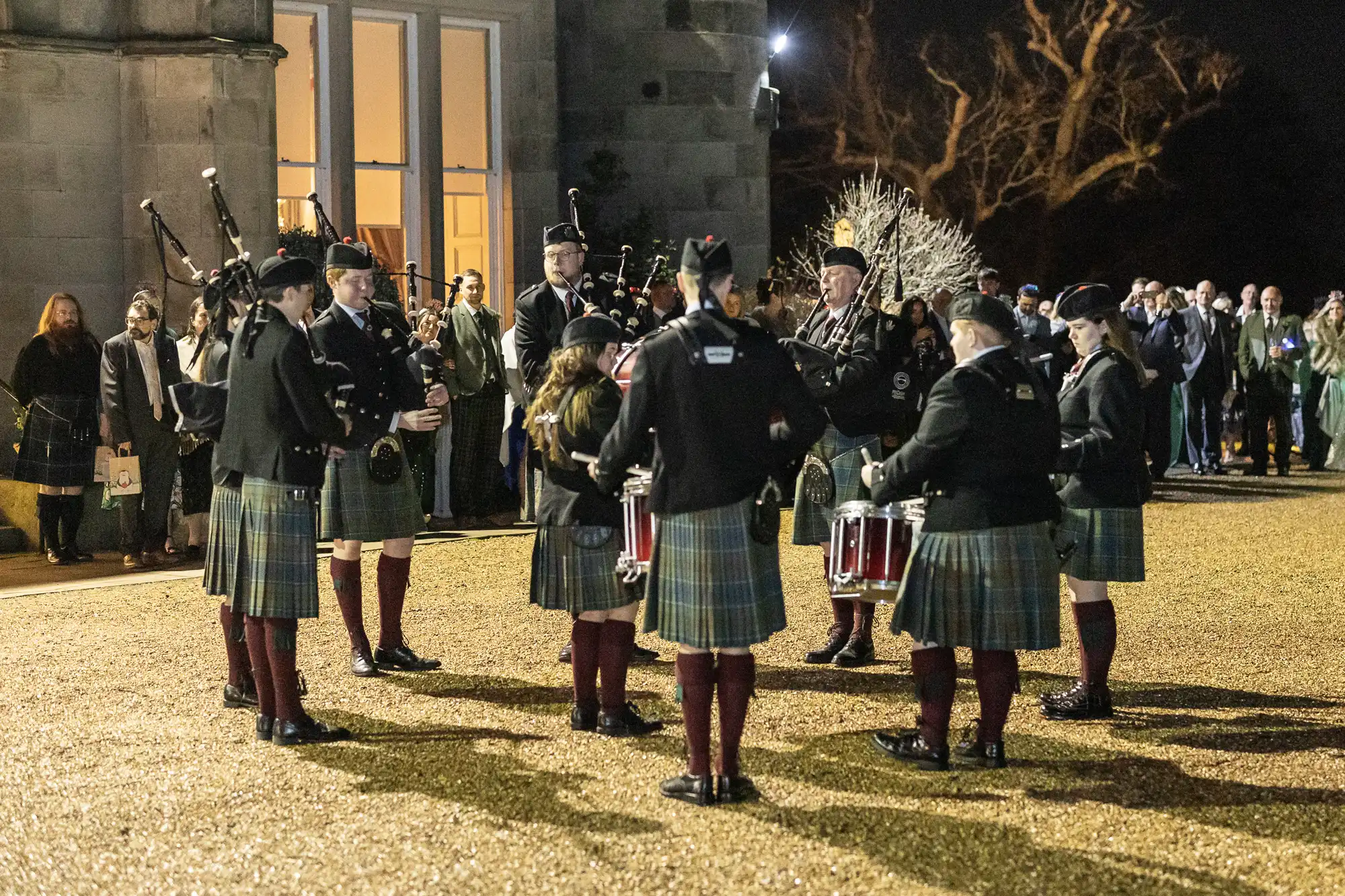 A group of people in traditional Scottish attire play bagpipes and drums outside a building at night, with a crowd of onlookers watching.