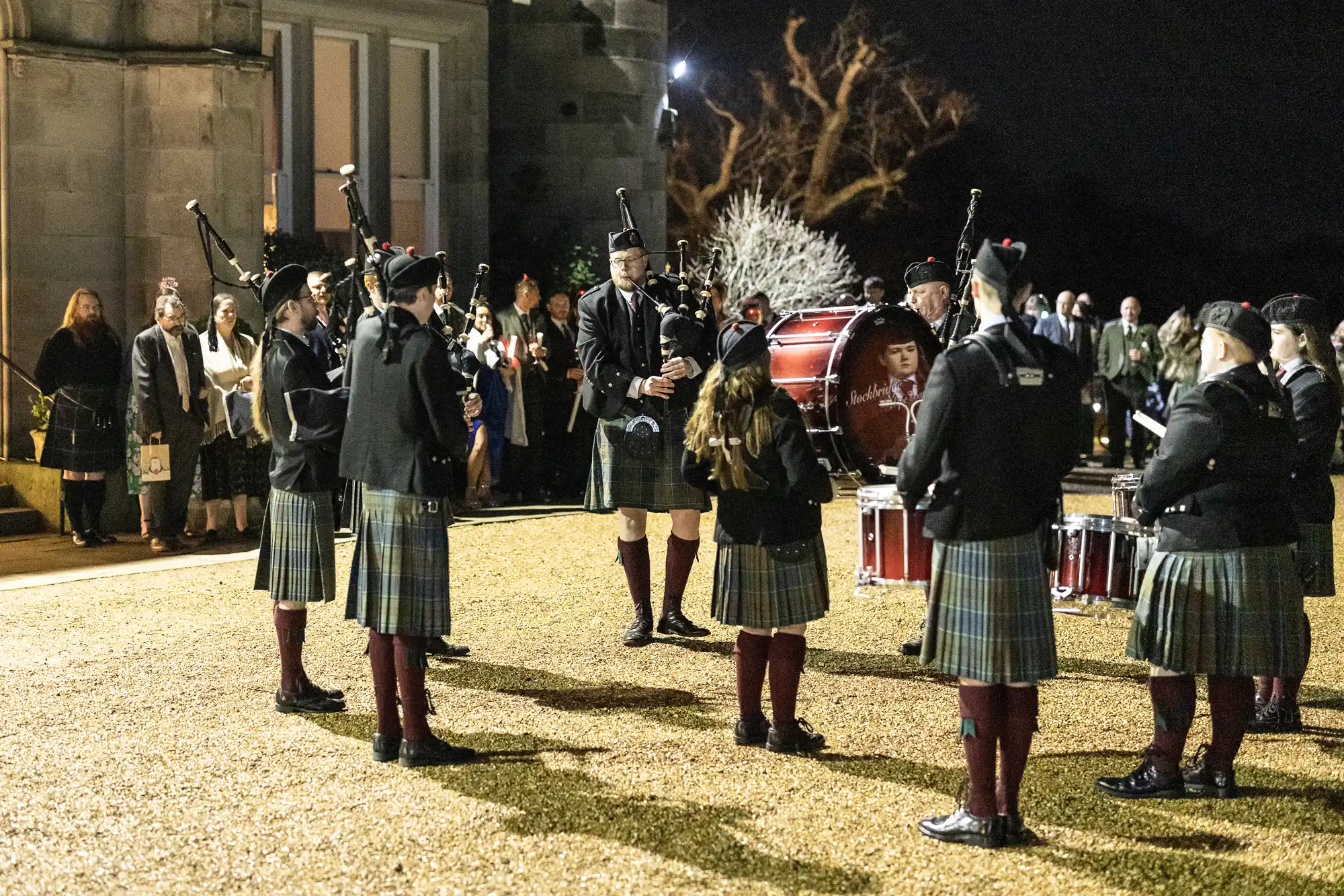 A Scottish pipe band, dressed in traditional attire, performs at night outside a building while a group of onlookers watches.