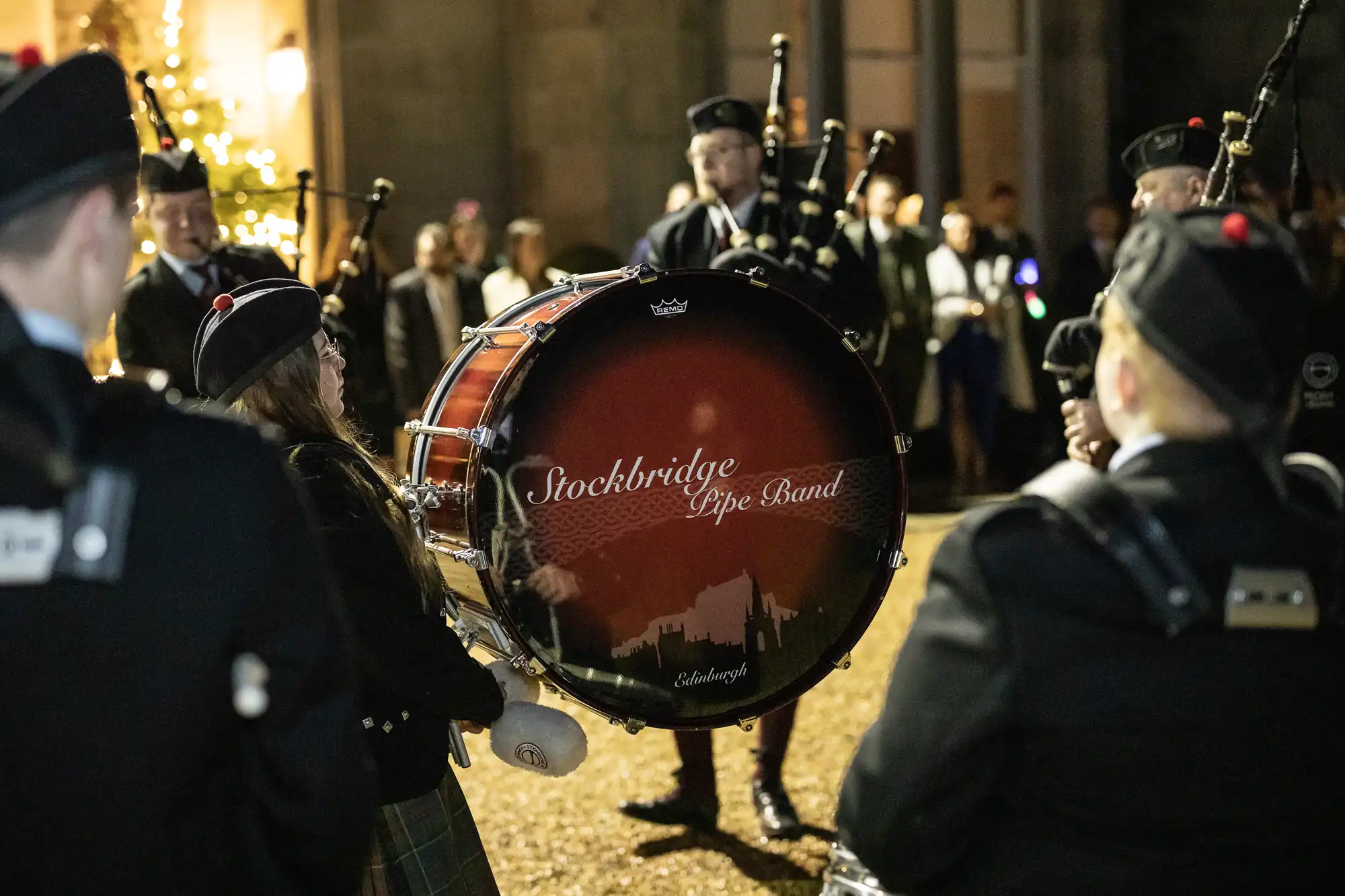 A drum with the text "Stockbridge Pipe Band" and musicians dressed in traditional attire performing outdoors at night.