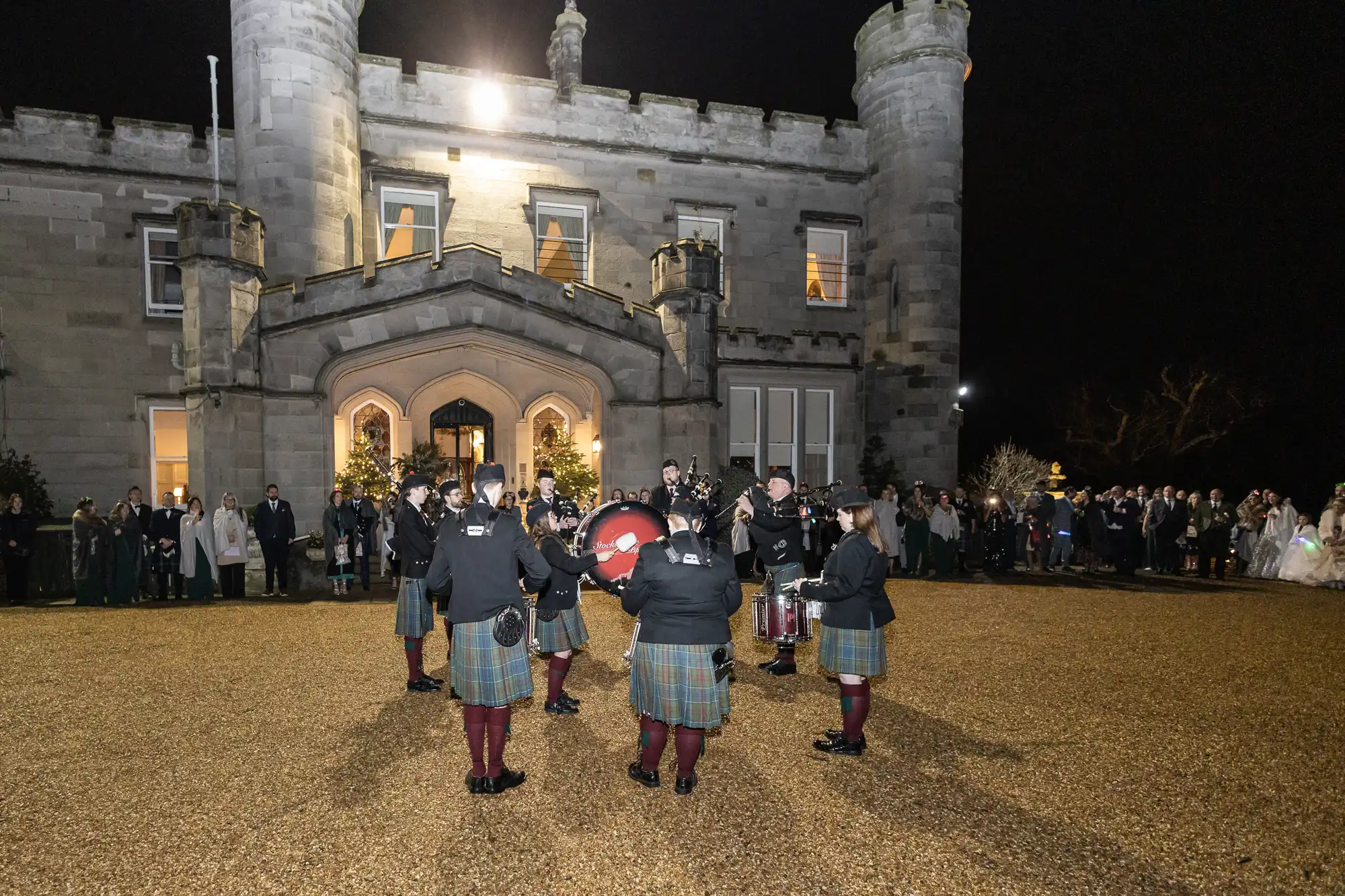 A small band wearing kilts performs outdoors at night in front of a large, illuminated stone castle, while a crowd of people watches from the sides.