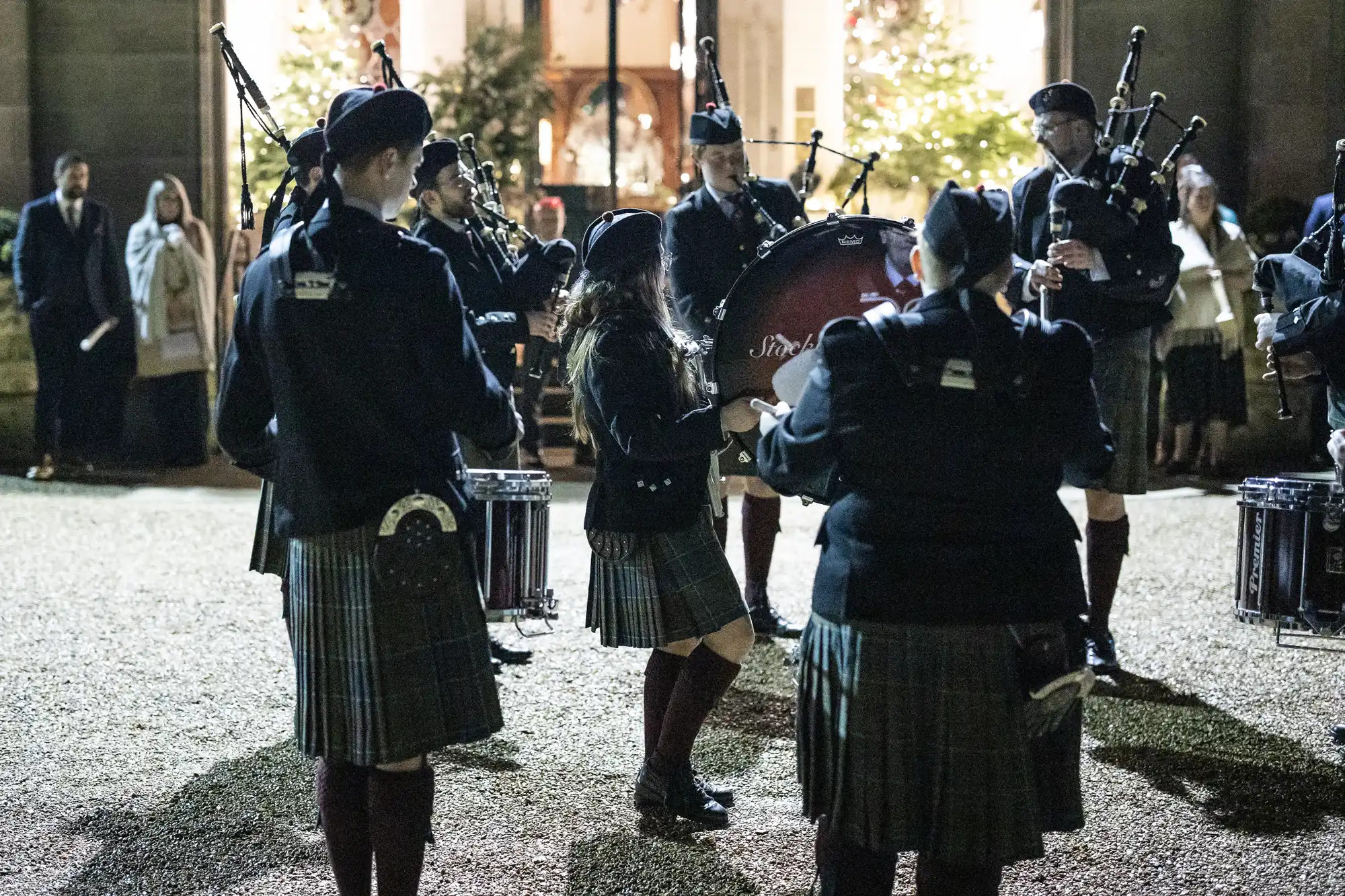 A group of people in traditional Scottish attire play bagpipes and drums outside a building with lit Christmas trees in the background.