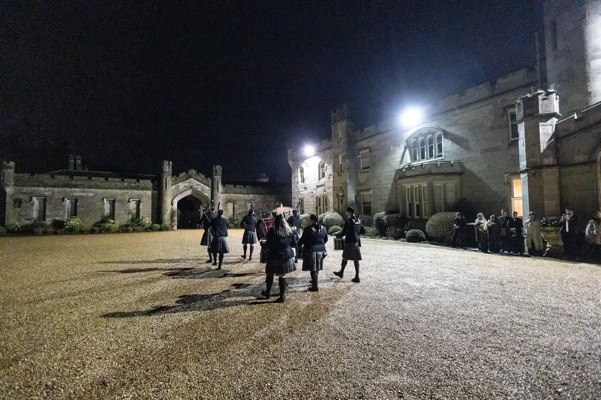 Group of people in traditional attire stand in a lit courtyard of a large stone building at night, with bystanders watching from the side.