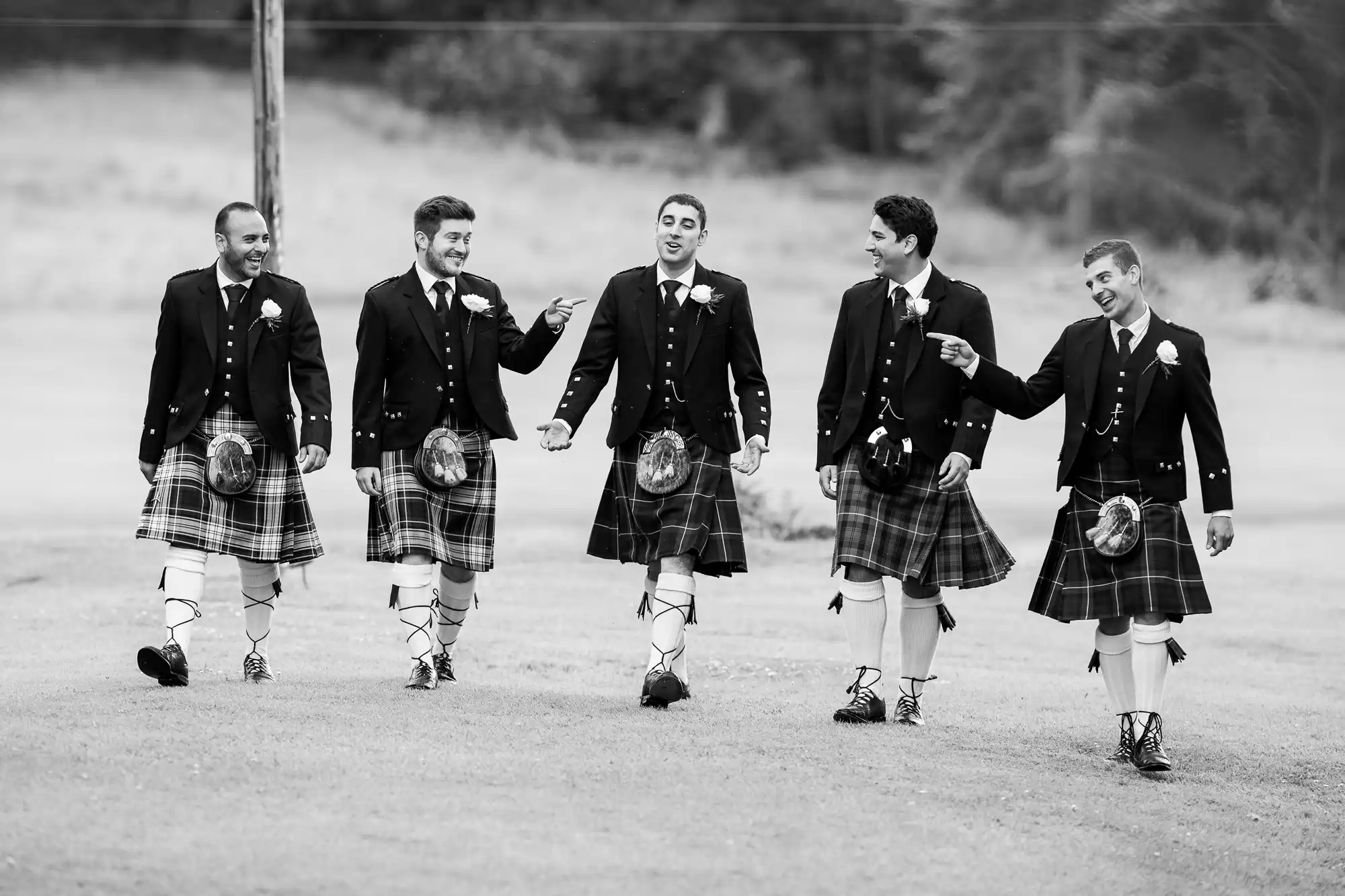 Groom and groomsmen wearing kilts and traditional Scottish attire walk and laugh together outdoors.