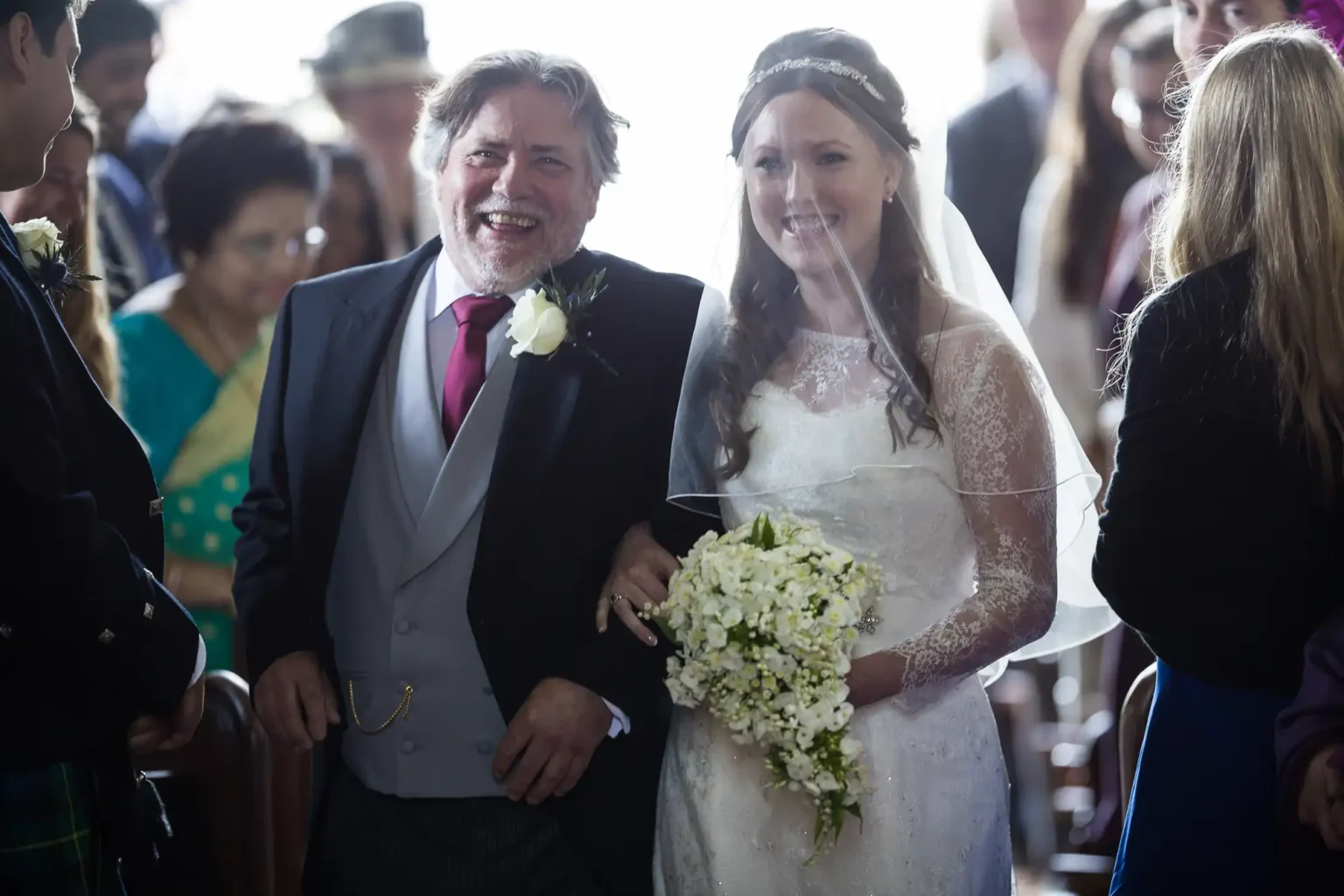 A bride in a white lace wedding dress and veil walks down the aisle arm-in-arm with her father in a suit, holding a white floral bouquet.