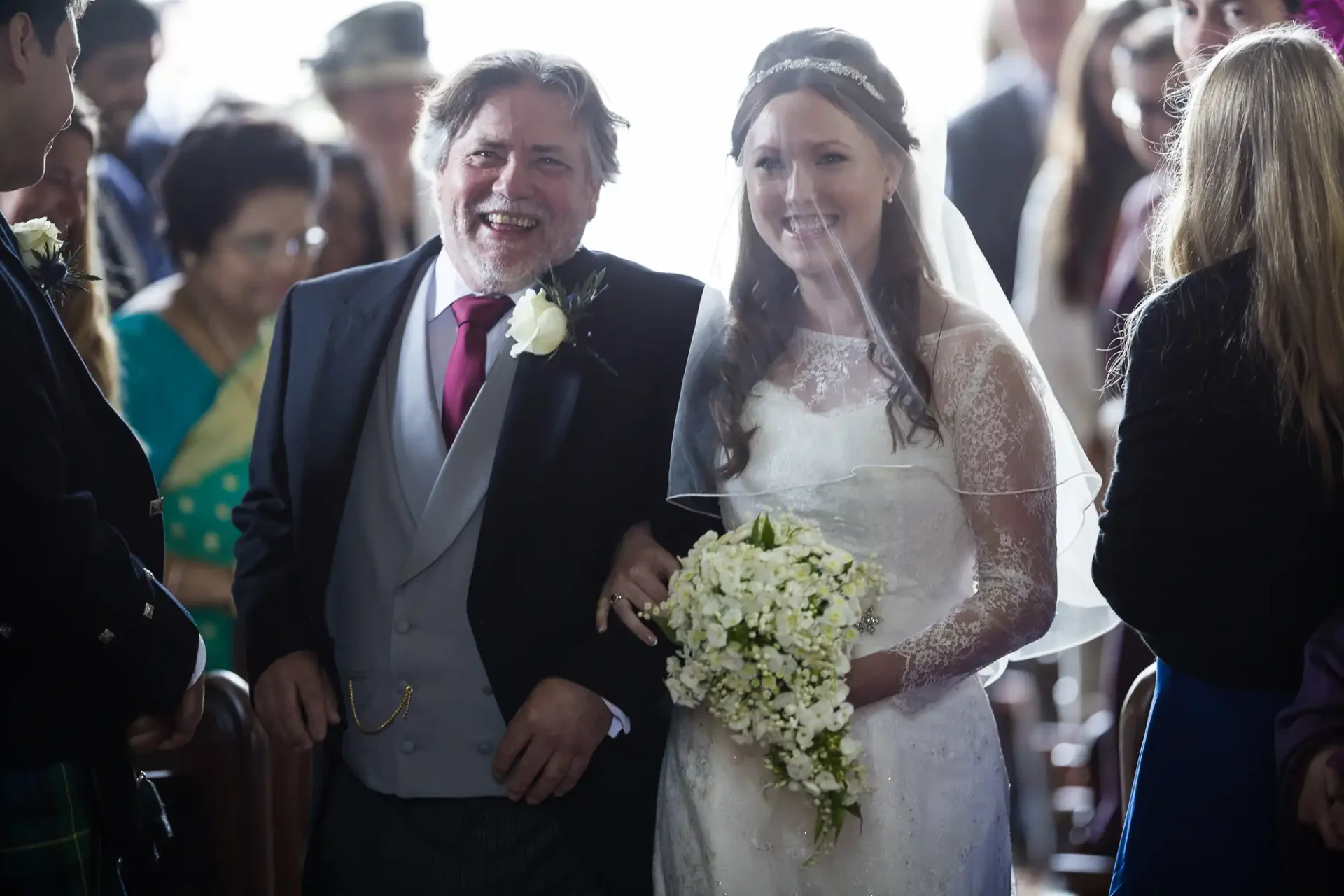 A bride in a white lace wedding dress and veil walks down the aisle arm-in-arm with her father in a suit, holding a white floral bouquet.