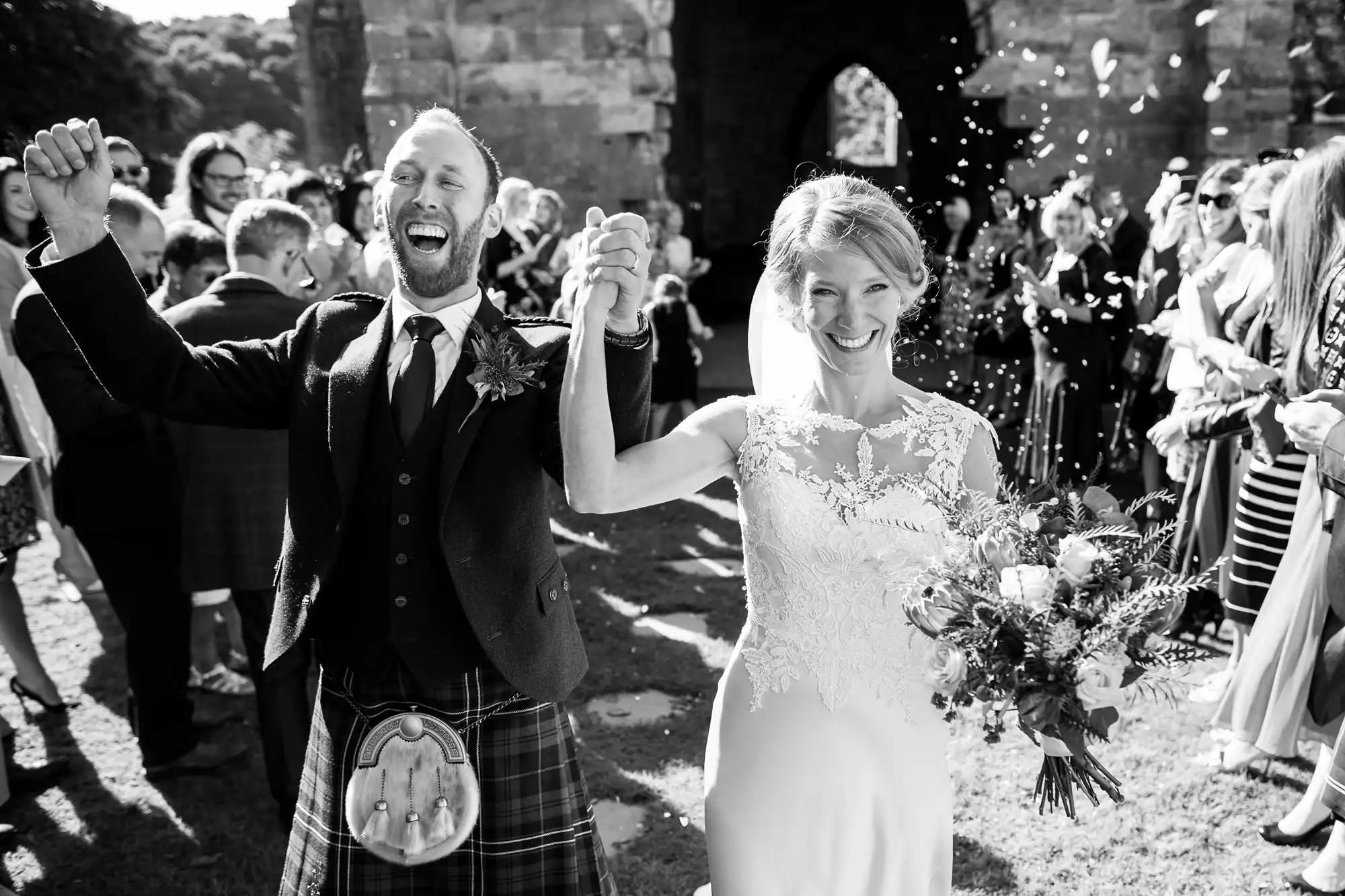A bride and groom, smiling and holding hands, walk down an outdoor aisle while guests cheer and throw confetti. The groom is wearing a kilt, and the bride is holding a bouquet.