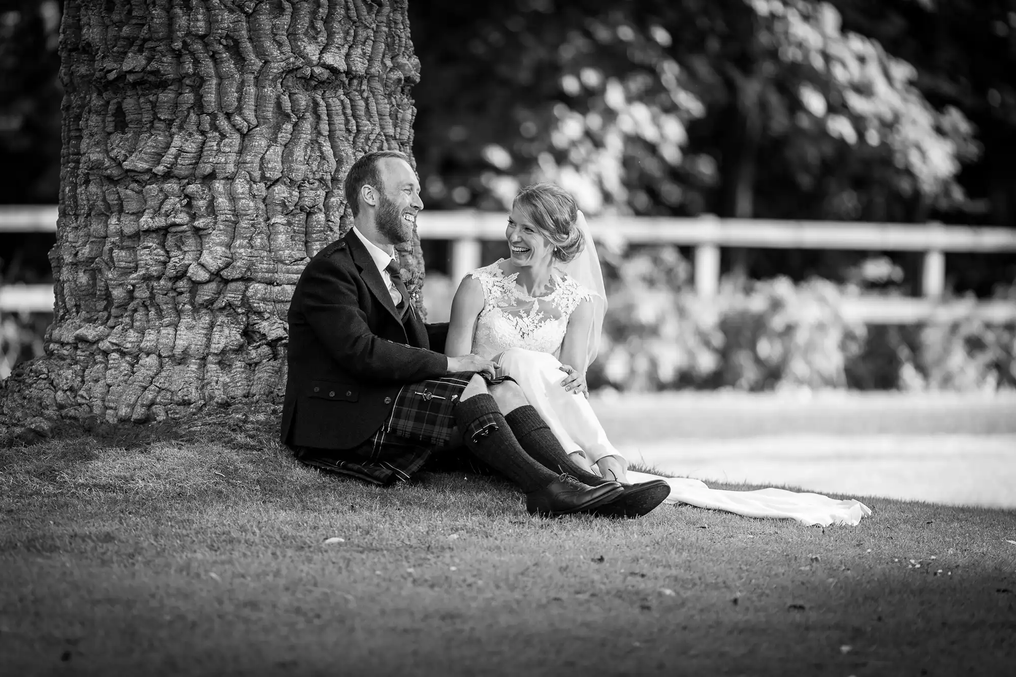 A couple in wedding attire sits on the grass, leaned against a tree, laughing and looking at each other.