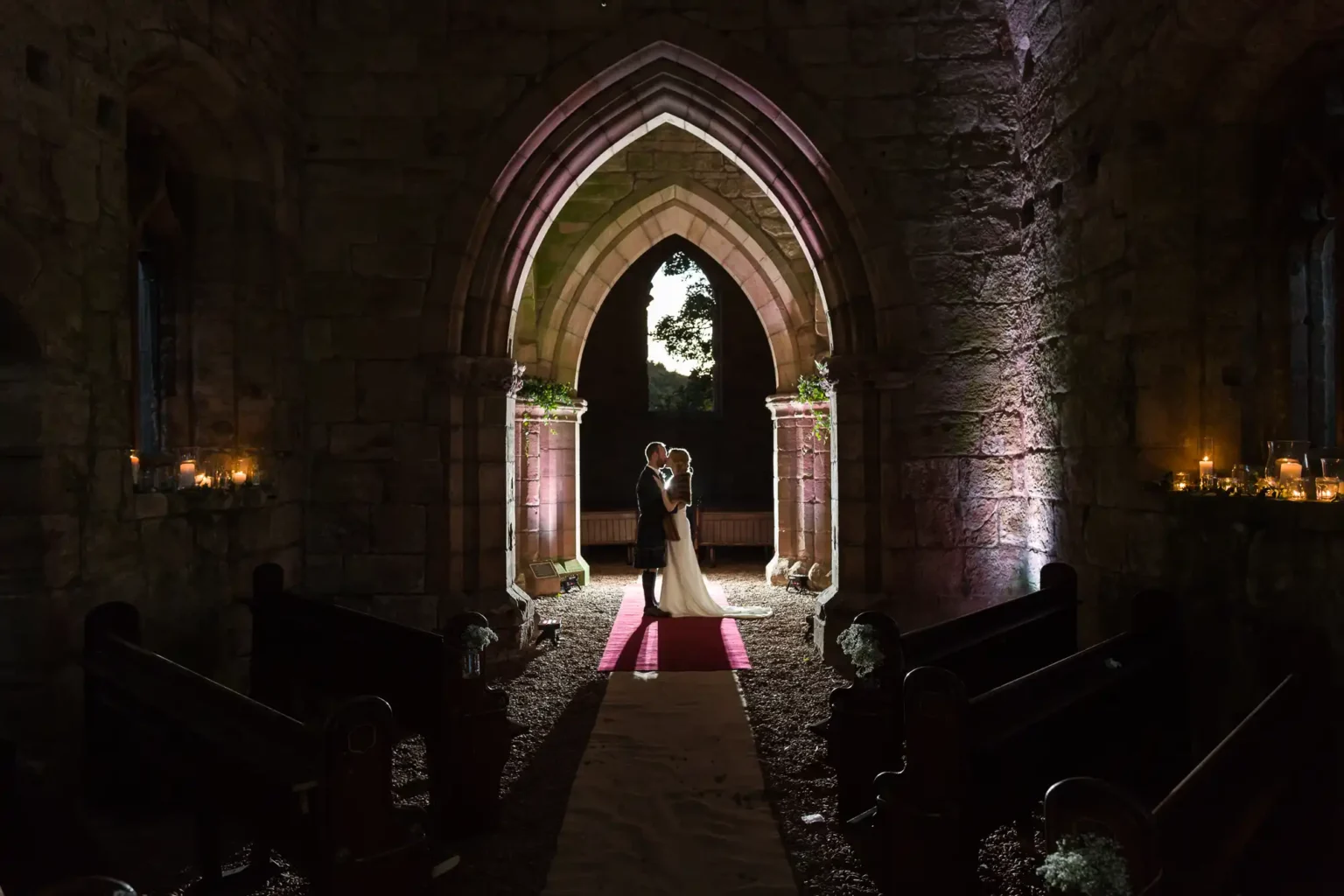 A newlywed couple shares a quiet embrace under a stone archway in a dimly lit church interior, with candles illuminating the space around them, at Dunglass Estate, East Lothian.
