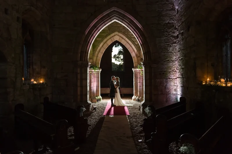 A newlywed couple shares a quiet embrace under a stone archway in a dimly lit church interior, with candles illuminating the space around them.