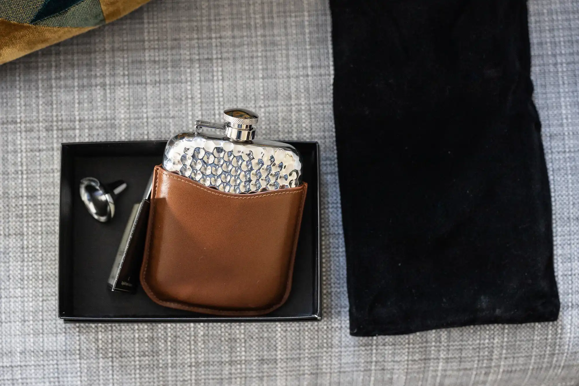 A metal flask with a brown leather cover, placed in a black box alongside a black drawstring bag and a small keychain. The items are on a gray fabric surface.