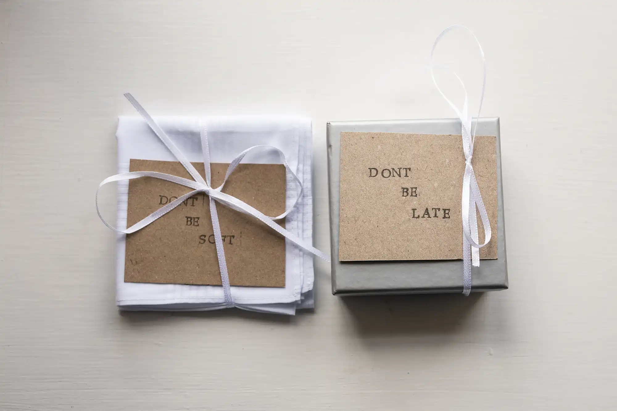 Two gift packages on a white surface: one contains fabric with a "DON'T BE SOFT" card, and the other is a box with a "DON'T BE LATE" card. Both are tied with white ribbons.