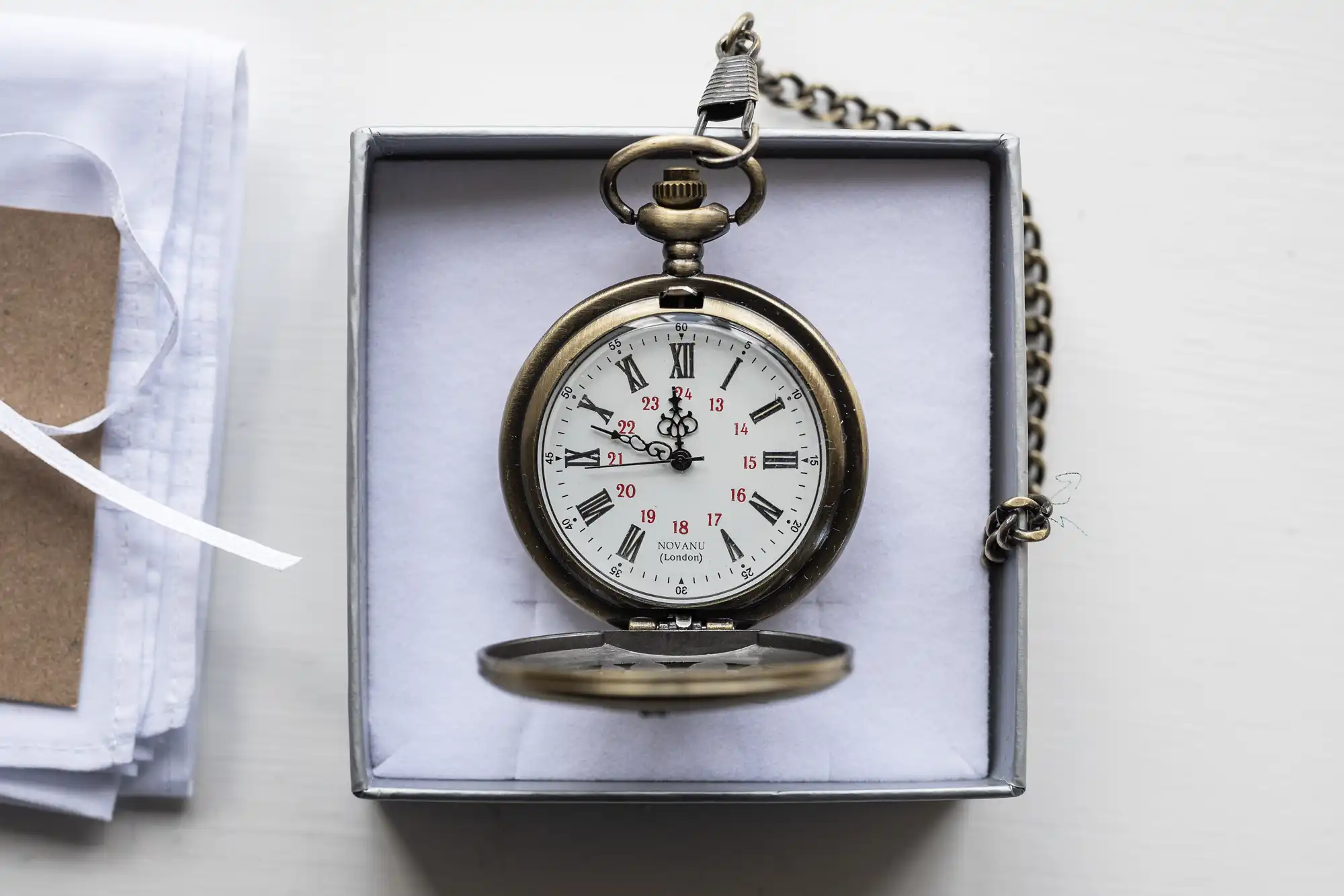 A vintage pocket watch with Roman numerals and a chain lies in an open box on a white surface, next to some folded papers.