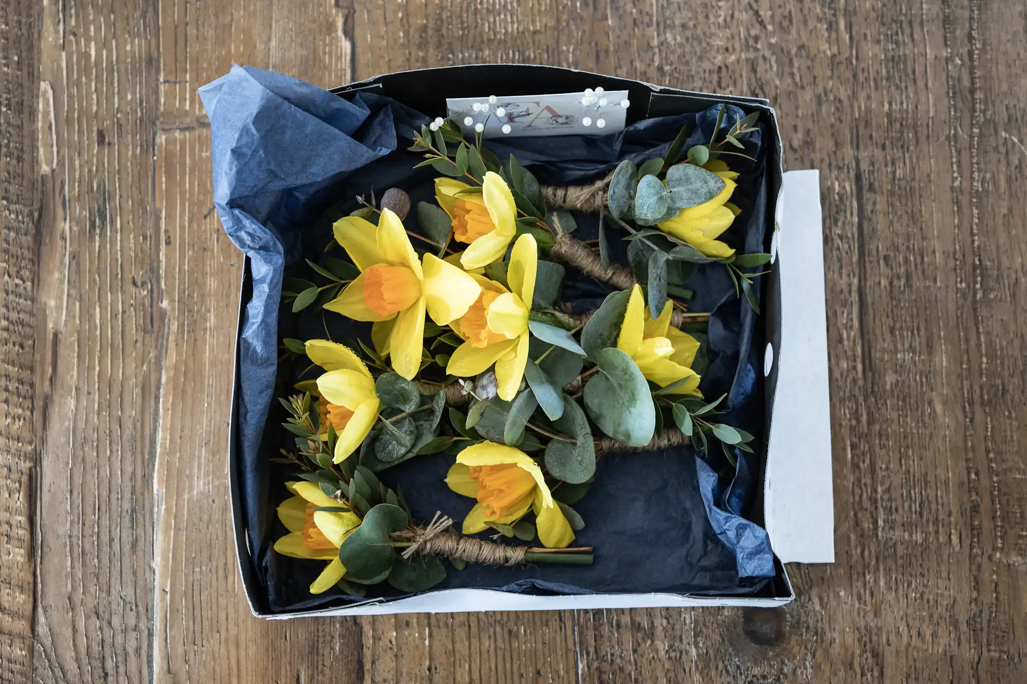 A box on a wooden table contains a neatly arranged assortment of yellow flowers, green leaves, and twine-wrapped stems.