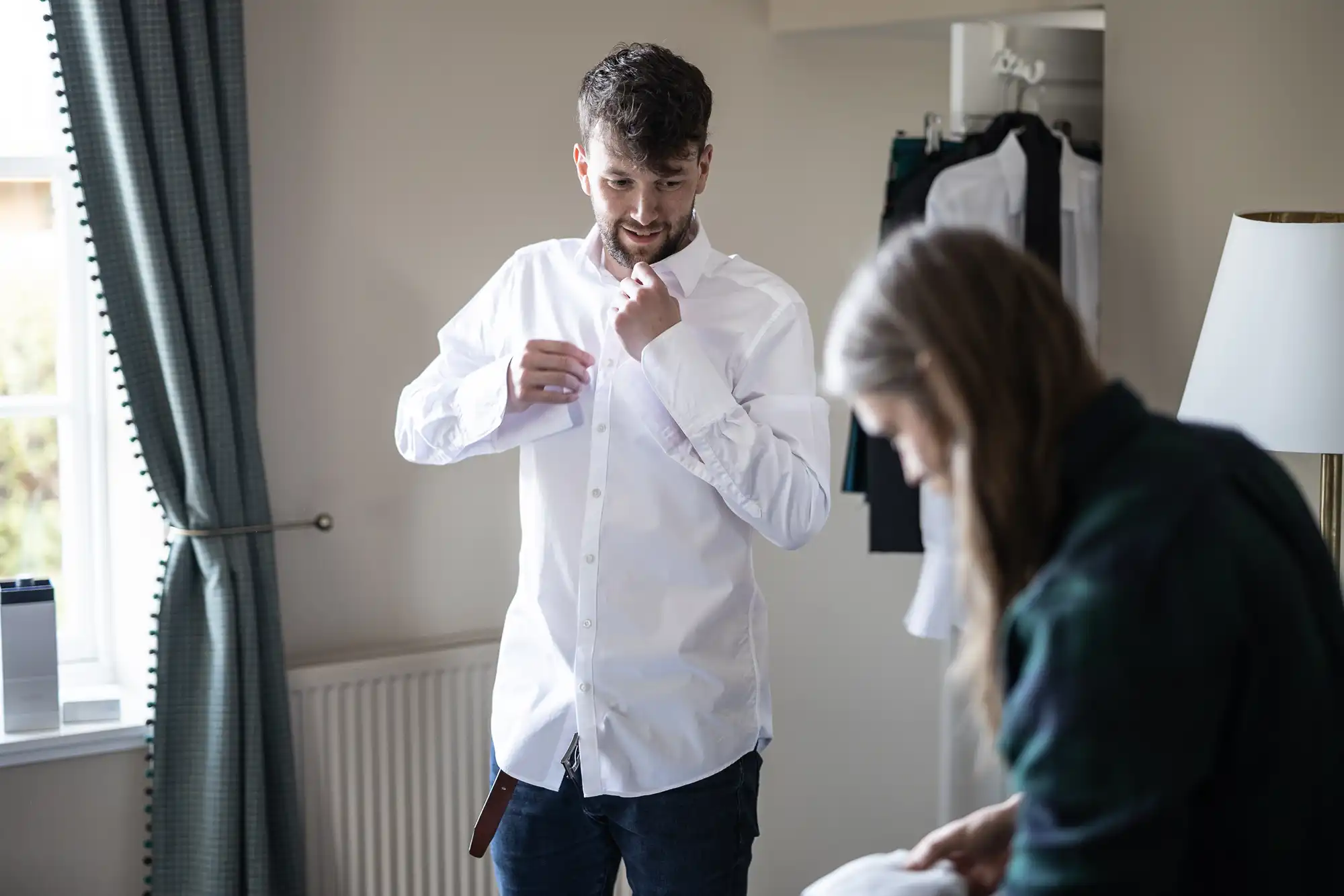 A man is buttoning up his white shirt in a room. Another person beside him is focused on an object in their hands. Clothing hangs in an open closet behind them.