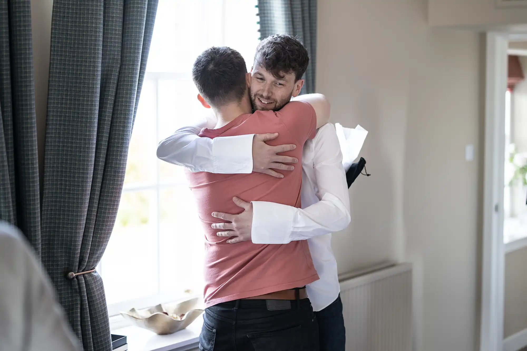 Two men hug near a window in a well-lit room with a radiator and grey curtains in the background. One man wears a white shirt, and the other wears a salmon-colored shirt.