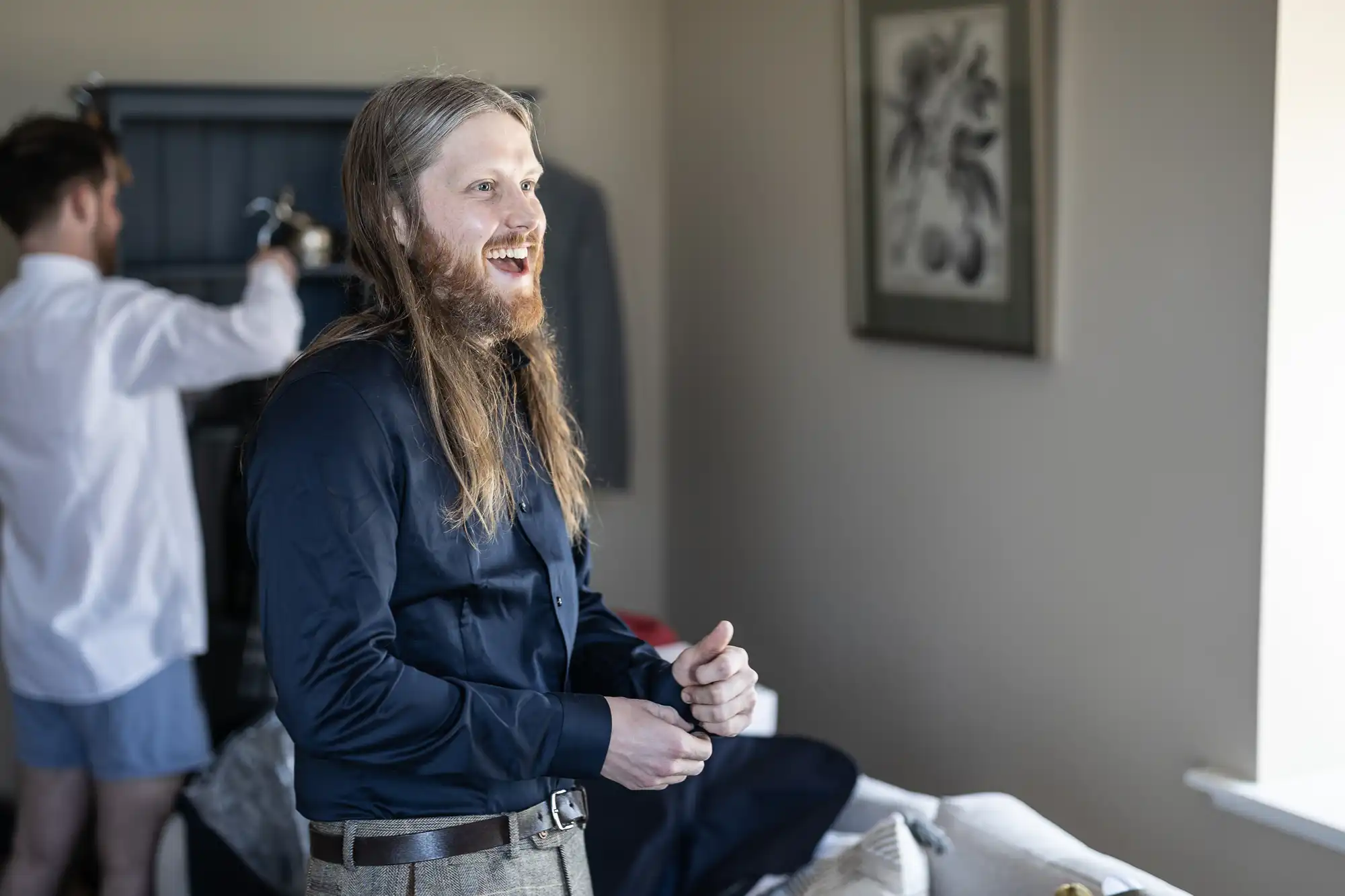 A man with long hair and a beard, wearing a navy shirt, is smiling and standing by a window. Another man in the background is adjusting a suit jacket on a hanger.