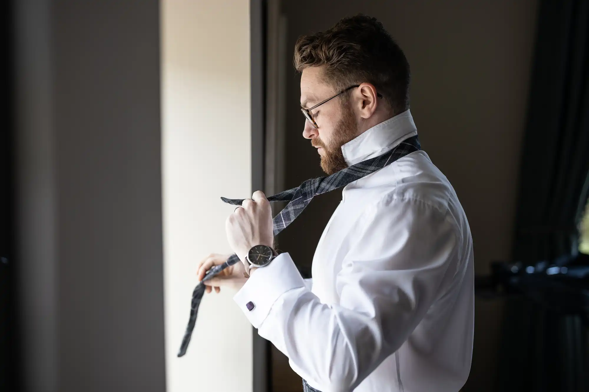 A man wearing glasses and a white shirt adjusts his tie while standing near a window.