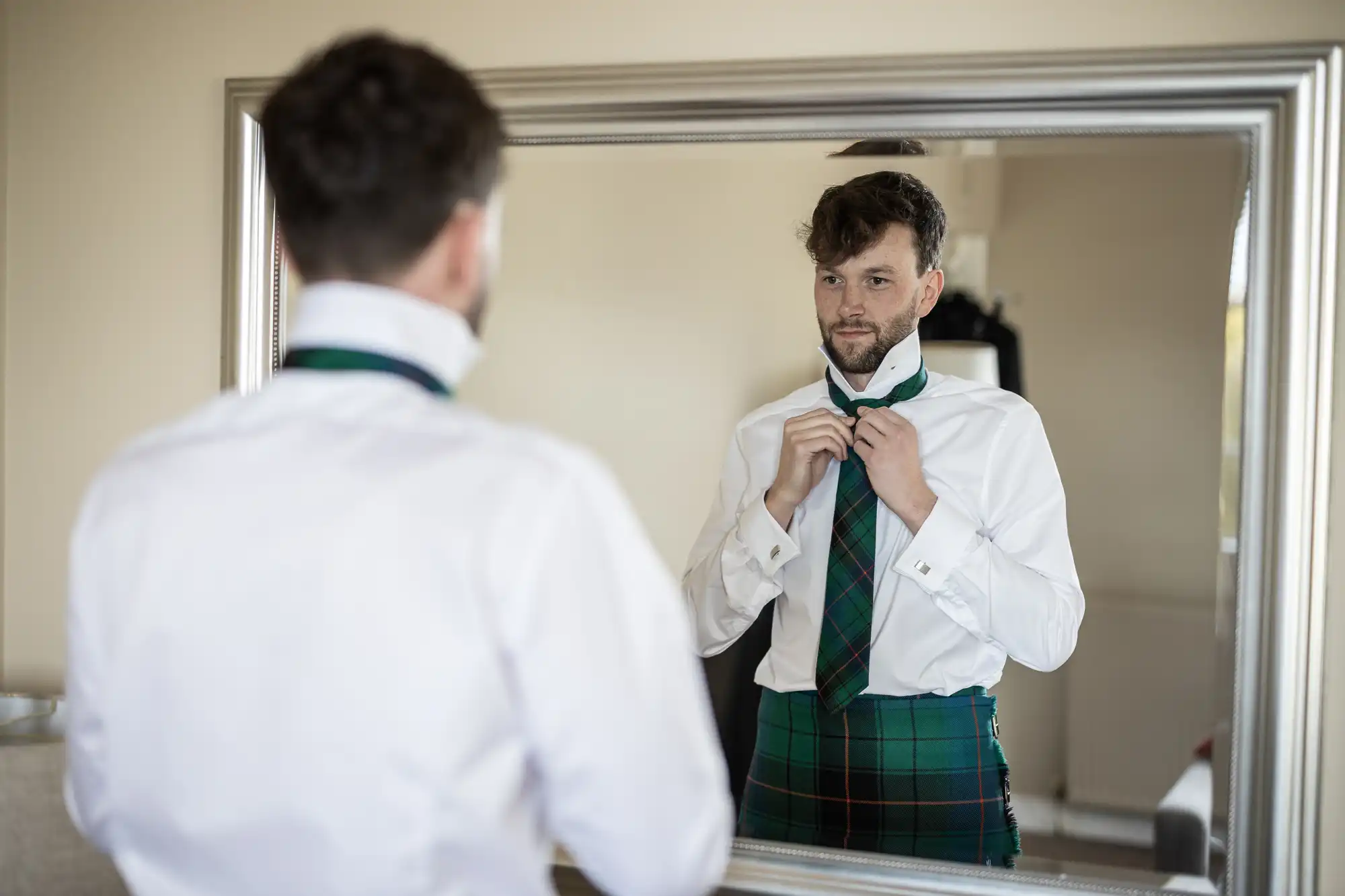 A man adjusts his necktie in front of a large mirror while wearing a white dress shirt and a green plaid kilt.