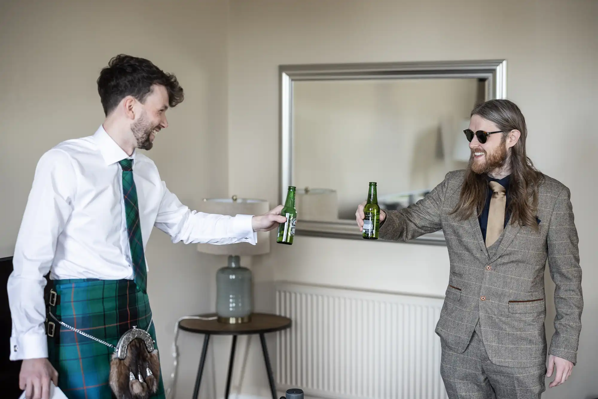 Two men indoors toasting with beer bottles. One man wears a kilt, shirt, and tie, the other wears a suit and sunglasses.