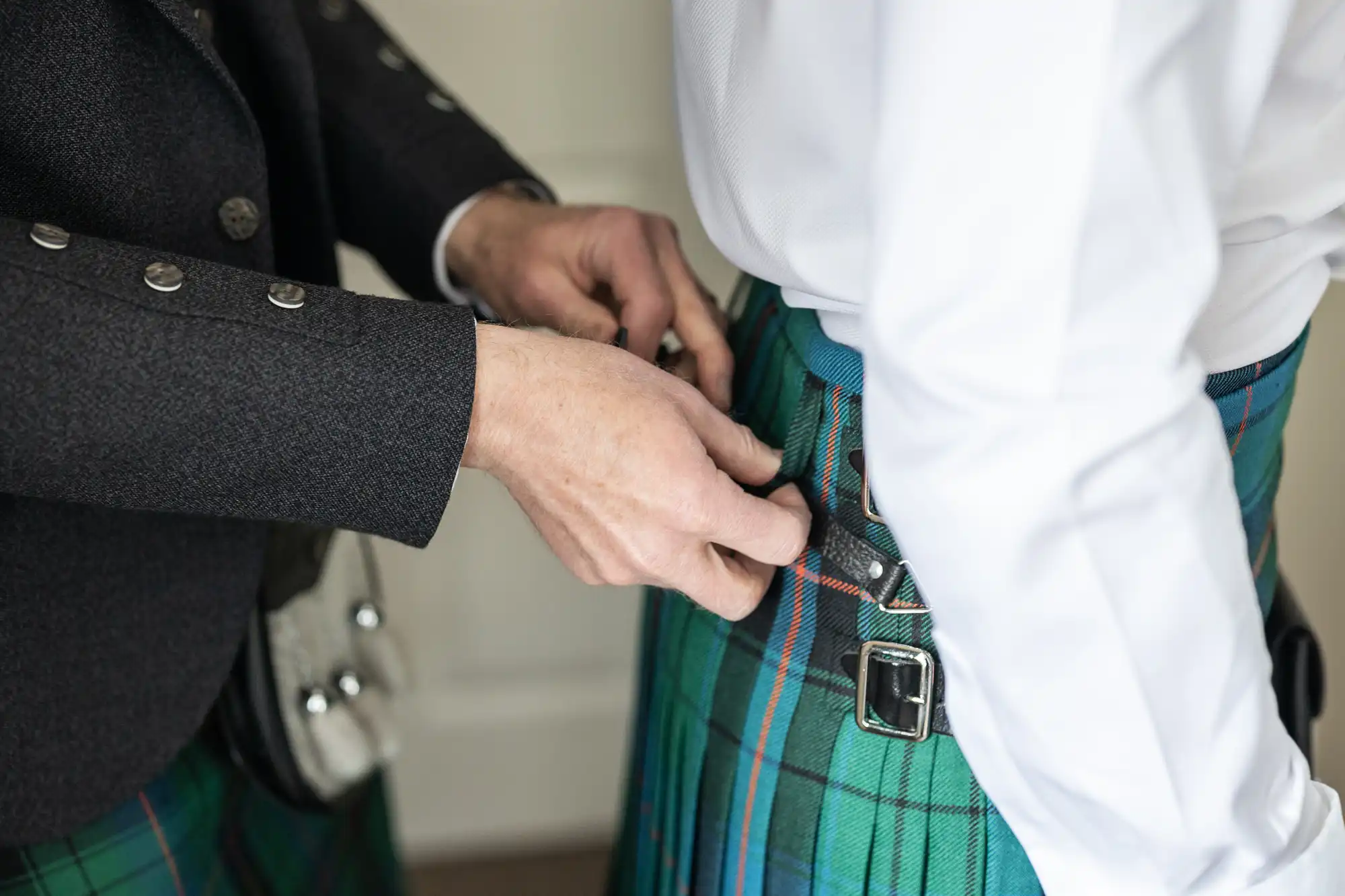 Person fastening a belt on another individual's green tartan kilt. Both appear to be wearing traditional Scottish attire.