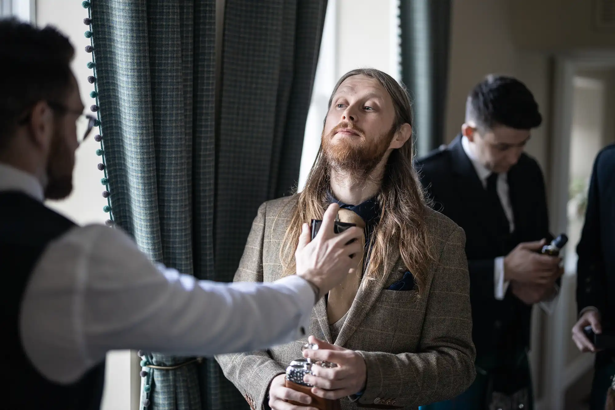 A man with long hair wearing a suit jacket is being sprayed with cologne by another man, while a third man is adjusting his tie in the background.