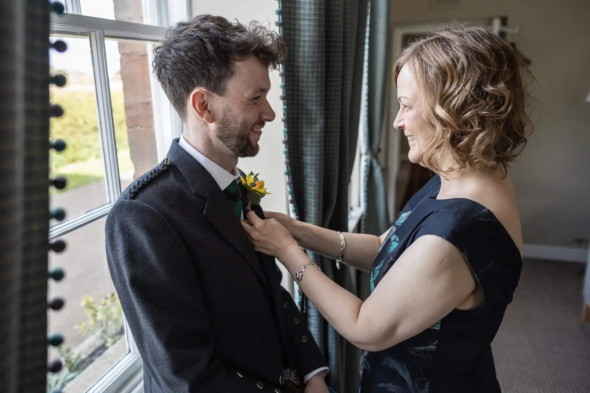 A woman adjusts the boutonnière on a man's lapel as they smile at each other indoors near a window.