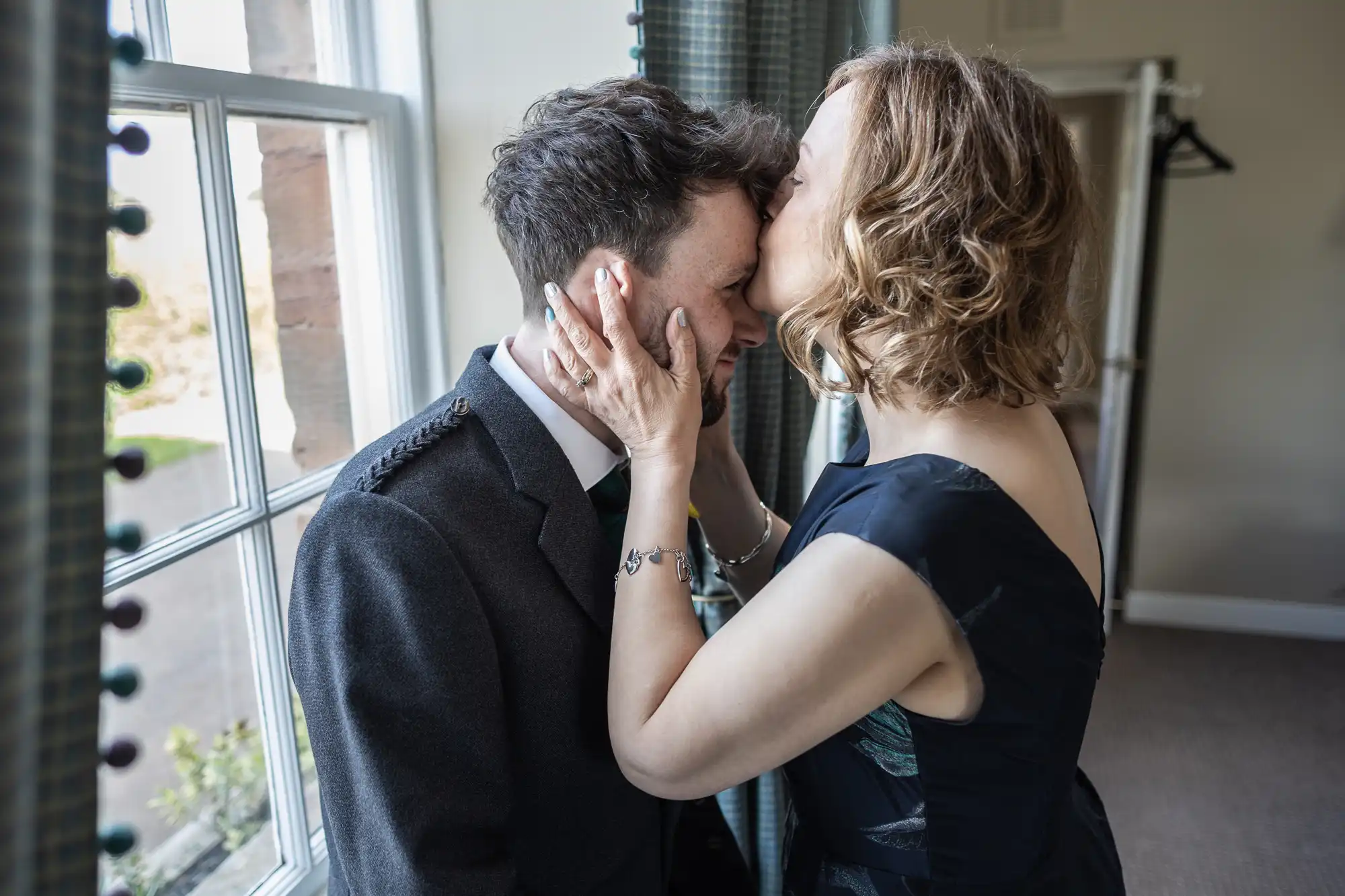 A woman in a dark dress is kissing a man in a suit on the forehead. They are standing near a window with green curtains in a well-lit room.