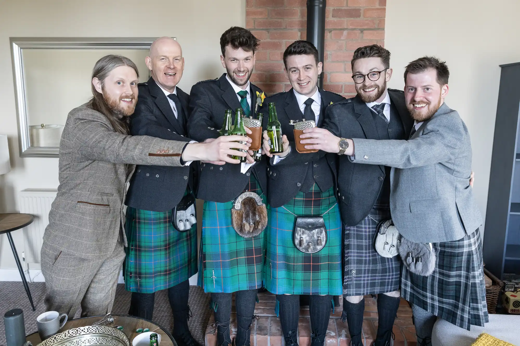 Six men, five wearing kilts and tweed jackets, and one in a checked suit, stand indoors and raise drinks in a toast.