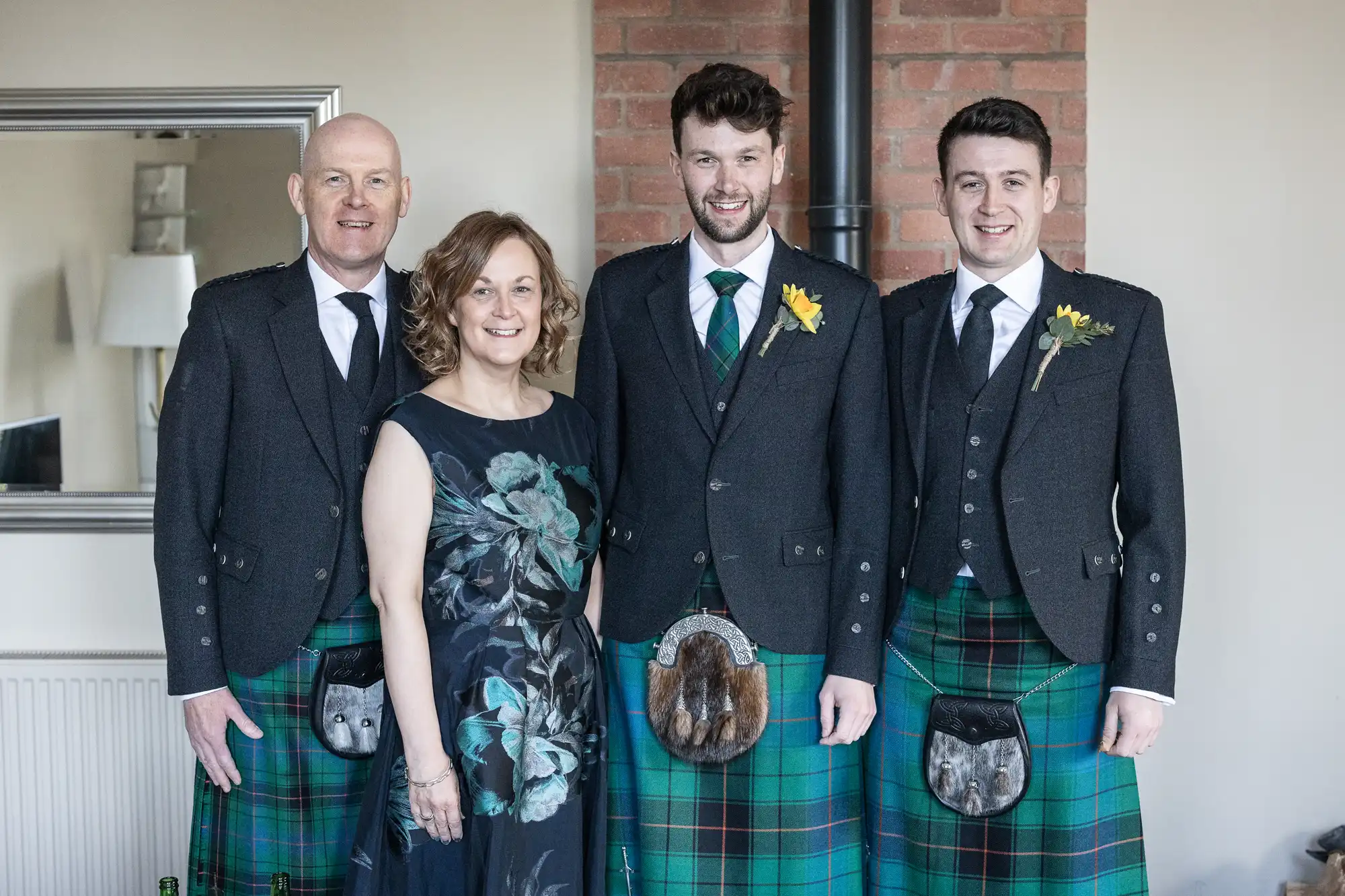 A group photo of three men in traditional Scottish attire and a woman in a black dress standing indoors. The men wear dark jackets and green tartan kilts with sporrans. The woman is smiling.