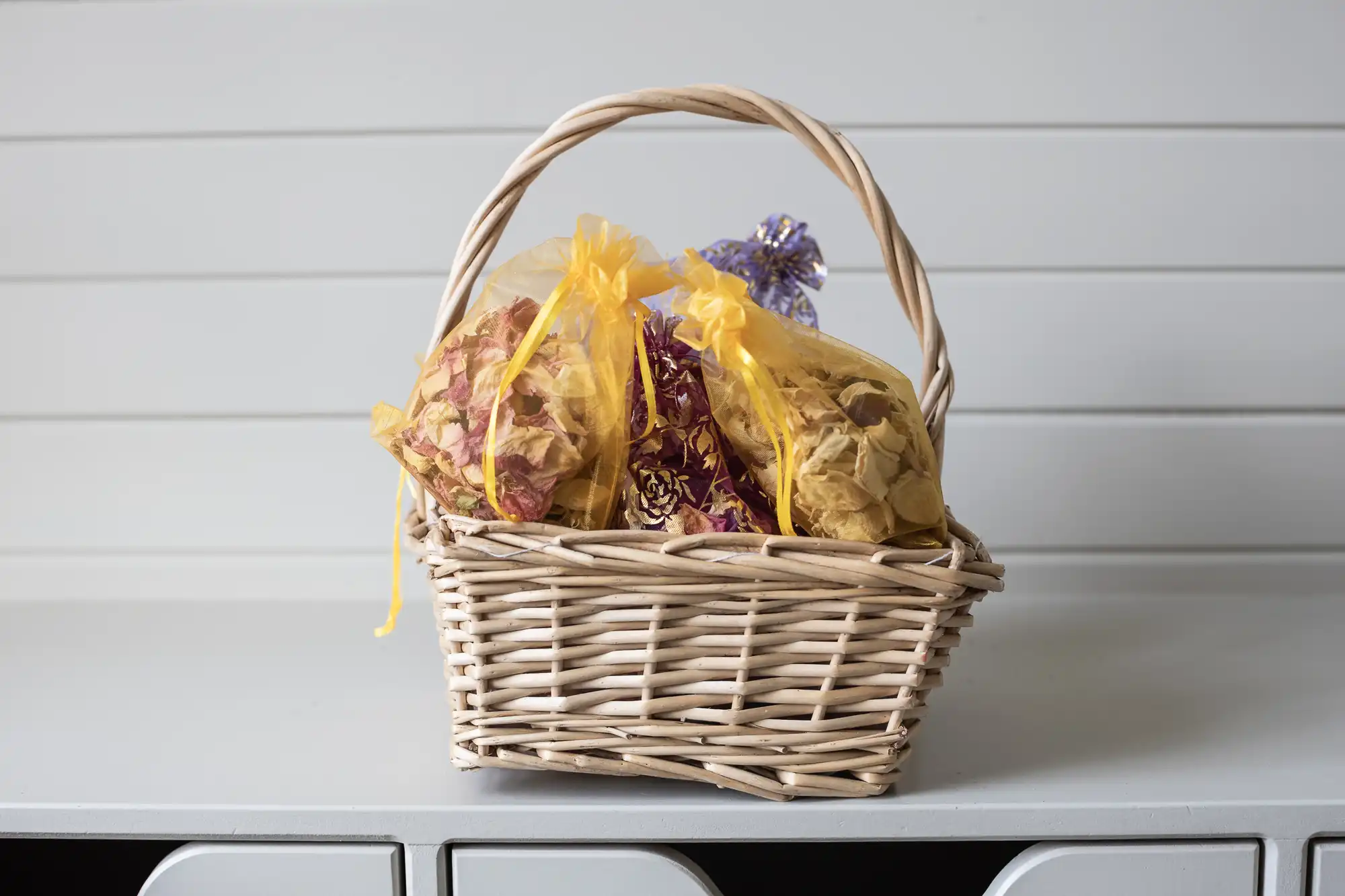 A wicker basket filled with small yellow and purple drawstring bags sits on a white surface with a light gray, paneled background.