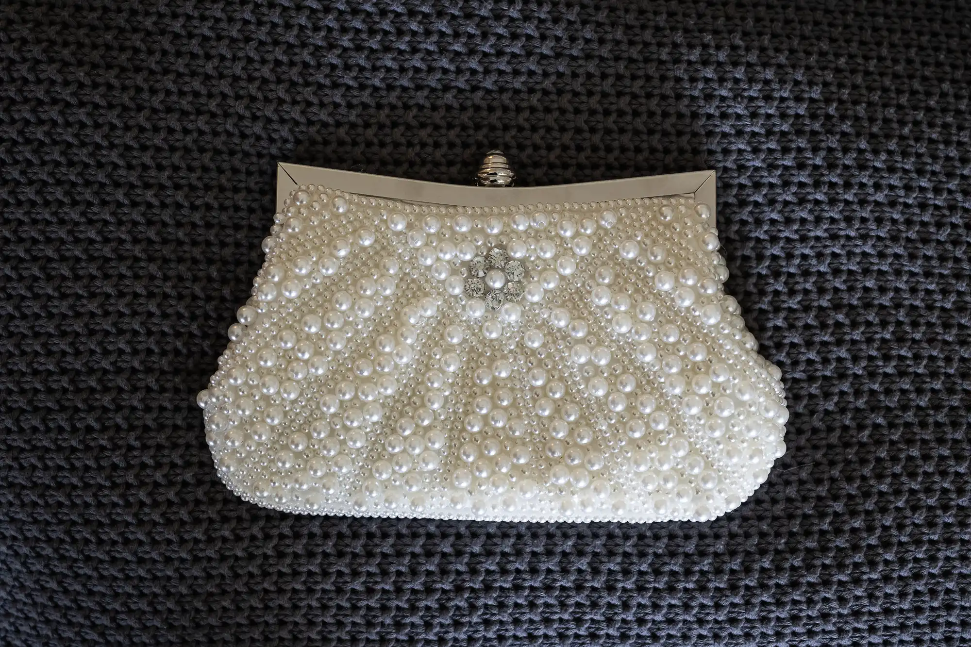 A small white clutch purse is adorned with numerous pearls, arranged in a symmetrical pattern, and features a silver clasp at the top. It is placed on a dark, textured surface.