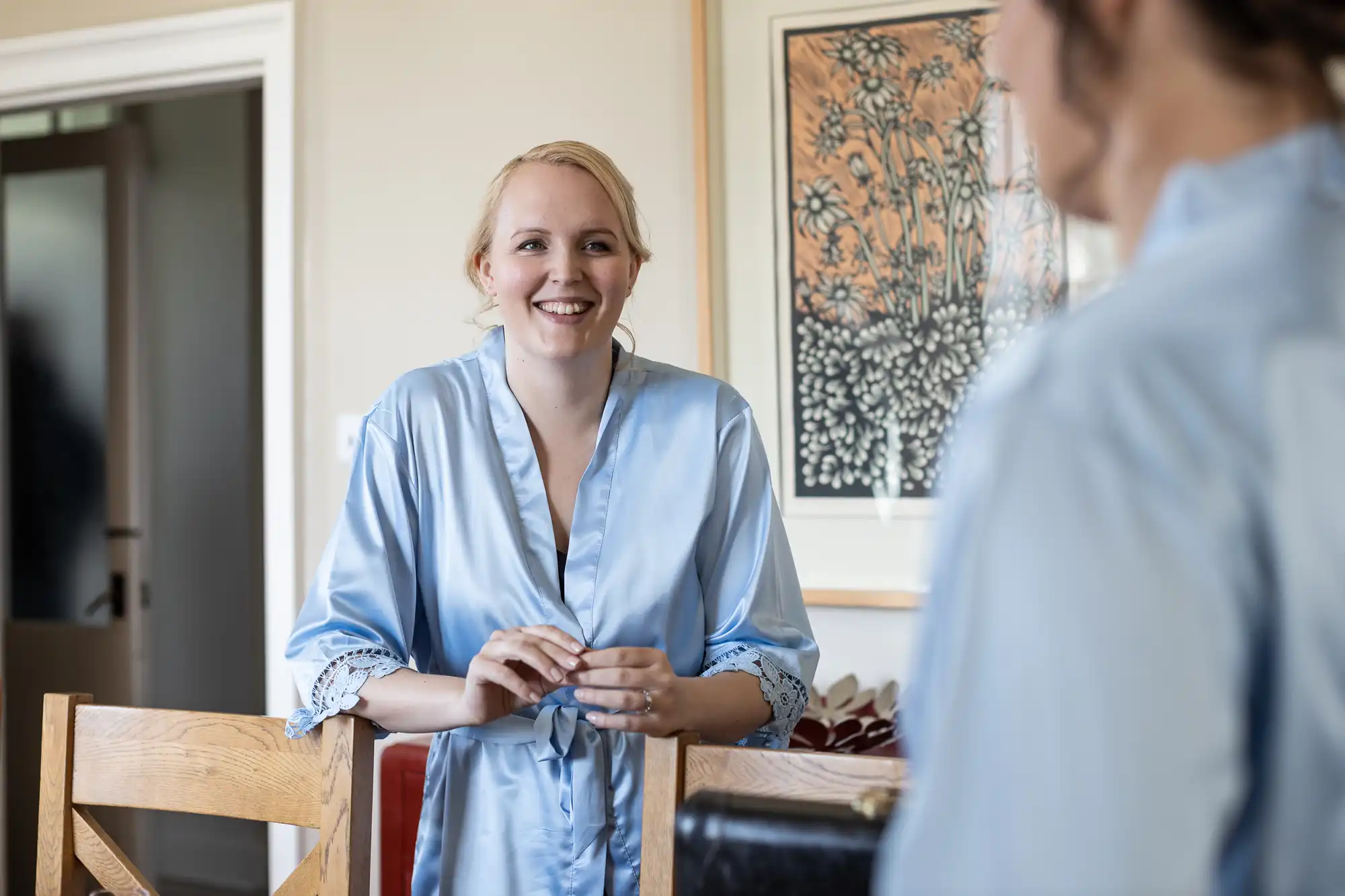 A woman in a light blue robe stands smiling near a wooden chair inside a room with art on the wall.