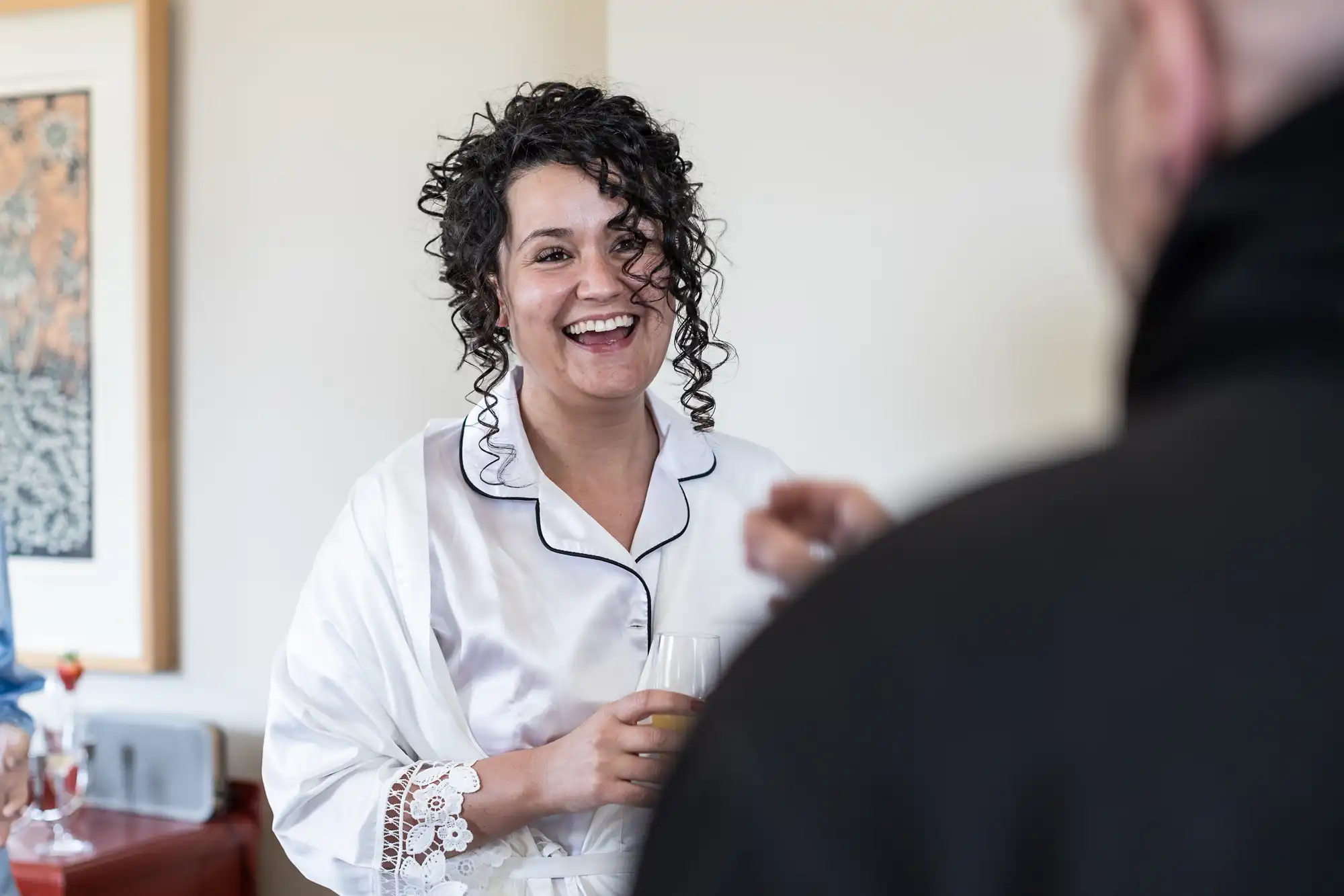 A woman with curly hair, wearing a white robe, smiles as she holds a glass and speaks with a person in the foreground.