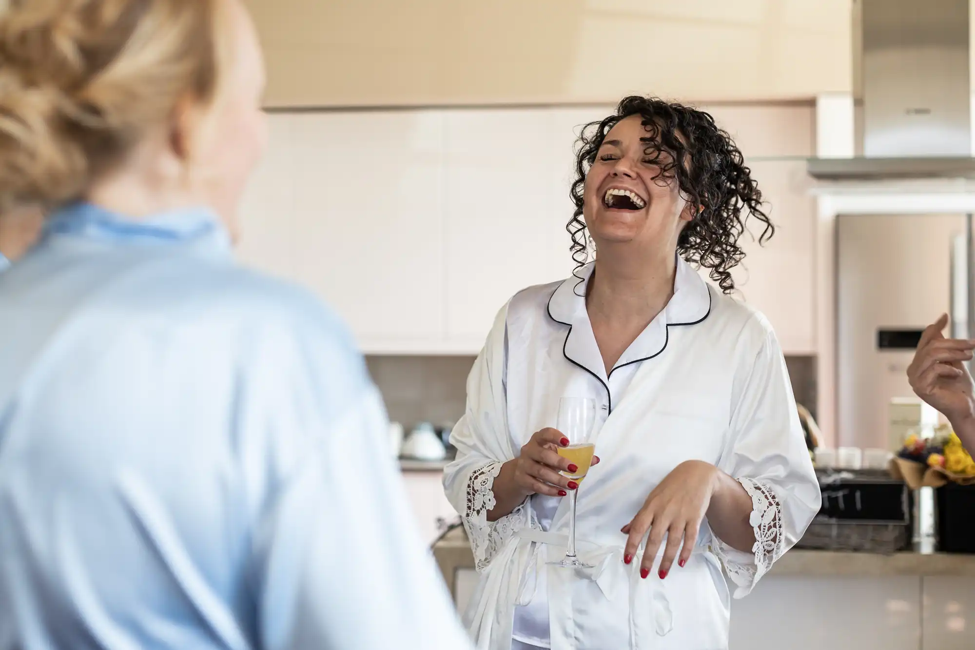 A woman in a white robe laughs and holds a glass of orange juice while talking with friends in a brightly lit kitchen.