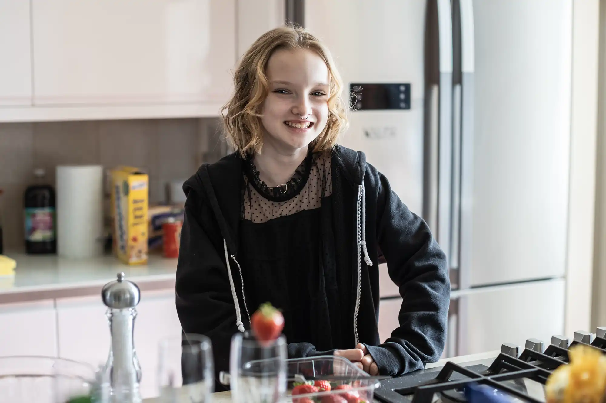 A young person with shoulder-length hair, wearing a black hoodie, stands smiling in a modern kitchen with various ingredients and utensils on the counter.